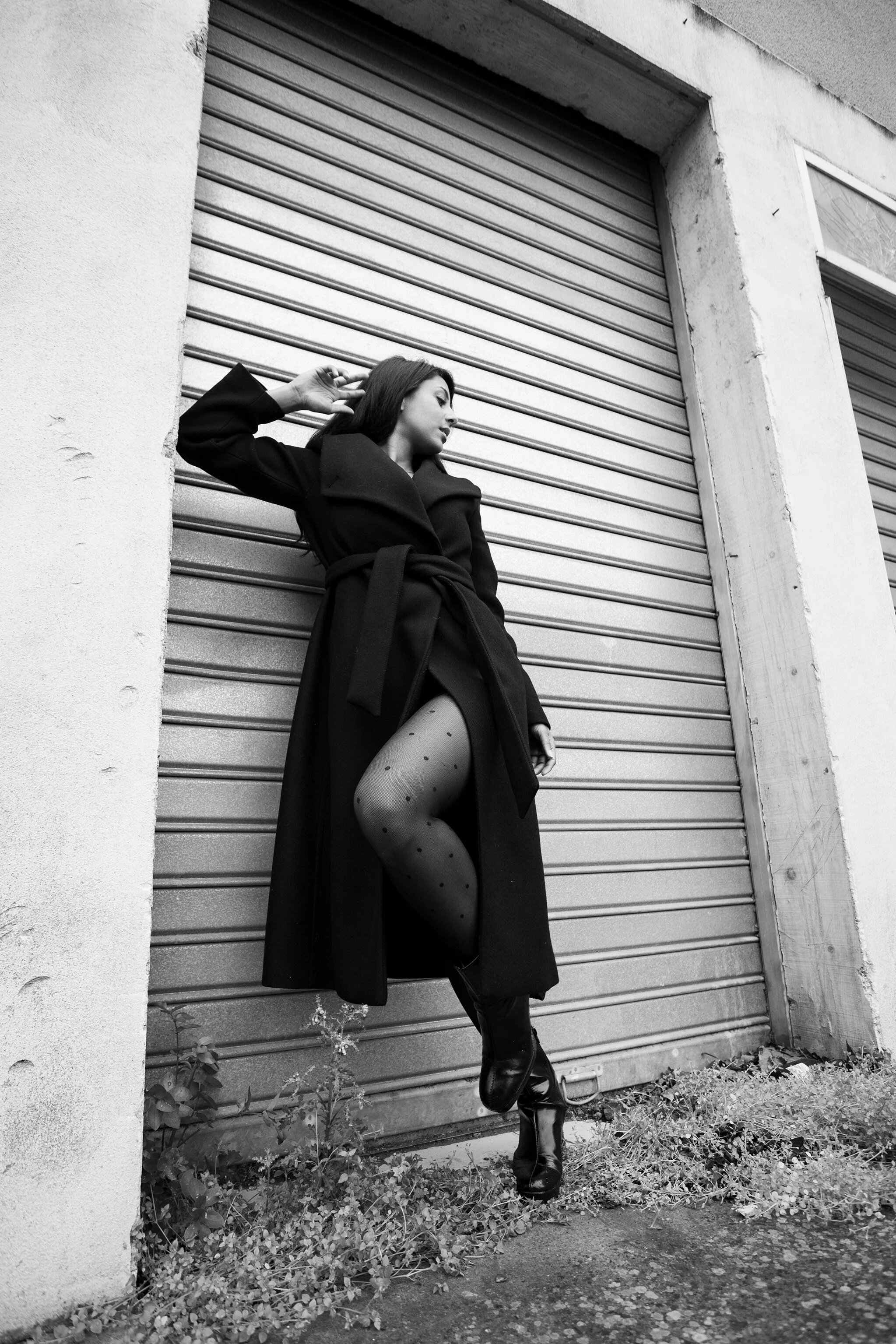 Femme posant contre un mur en béton avec une porte métallique à volet roulant, portant un manteau long, des collants à pois et des bottines en cuir.
