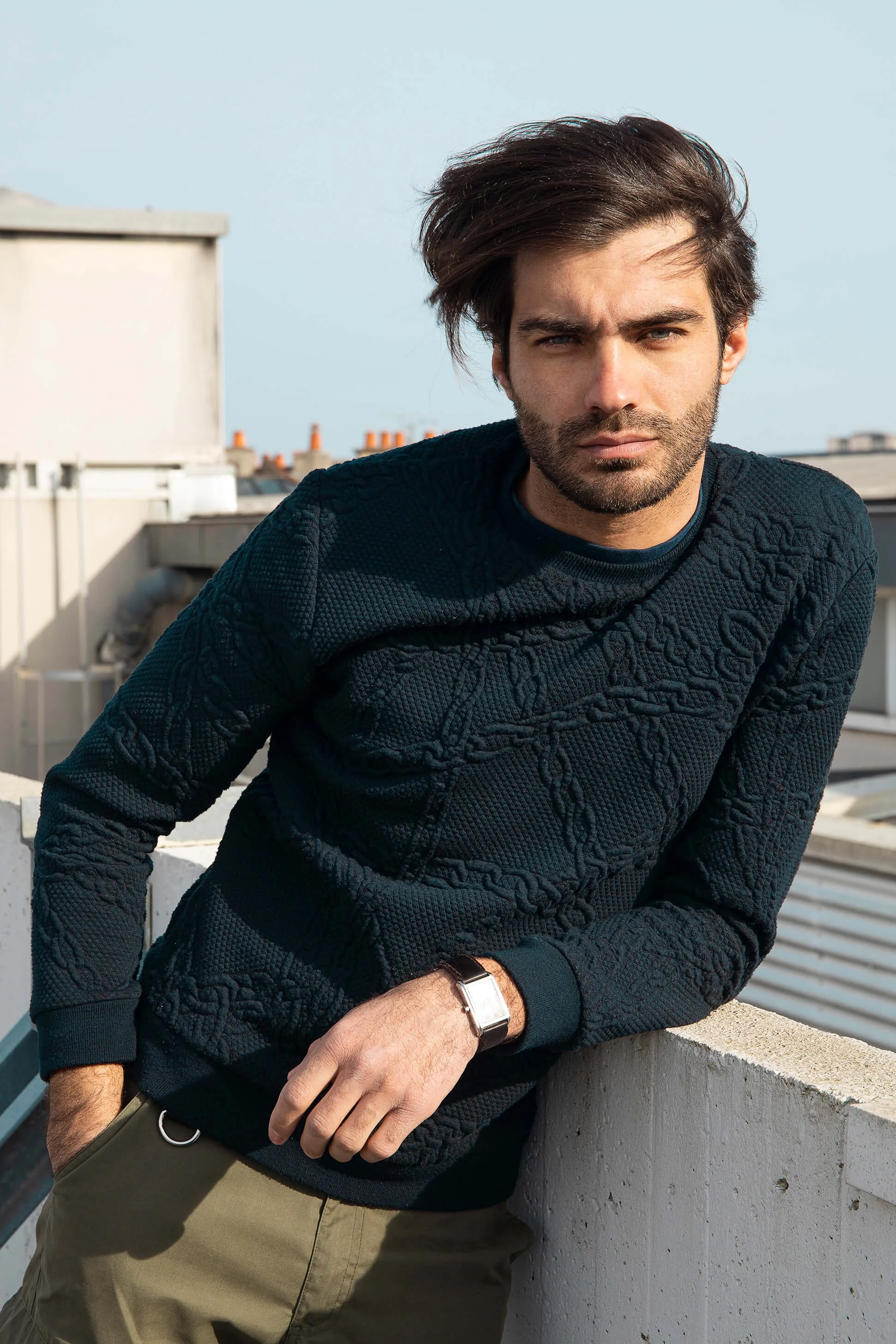 Jeune homme avec cheveux bruns, barbe, portant pull noir texturé et montre argentée, posé sur un balcon en ville, regardant l'objectif.