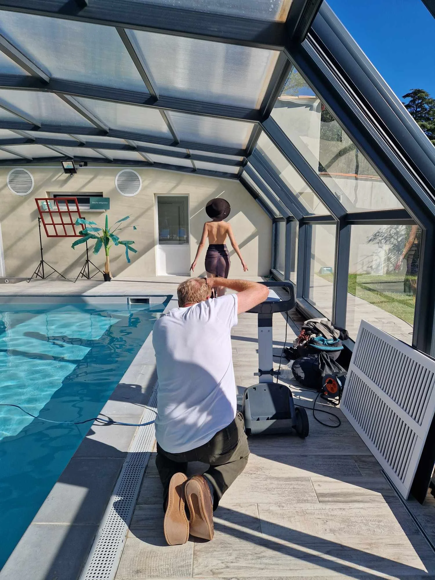 Portrait de François Fauré prend une photo d'une femme à côté d'une piscine à l'intérieur d'une serre ou véranda avec une structure en verre.