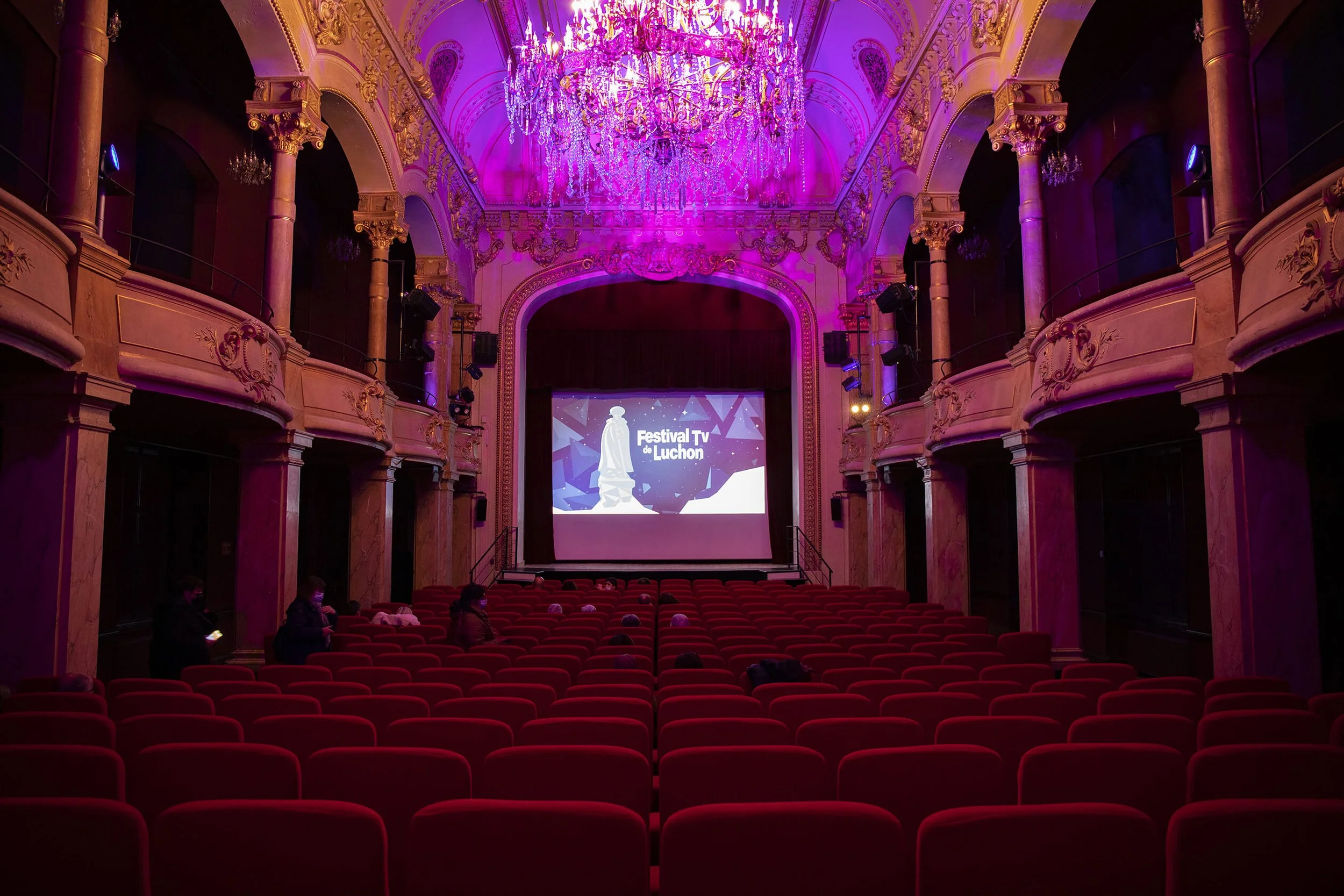 Décor élégant du théâtre à Luchon, mettant en valeur des détails architecturaux et un lustre scintillant. Ambiance chaleureuse du théâtre de Luchon, avec des couleurs vives et un éclairage tamisé. Événement au Festival TV de Luchon, avec une scène il