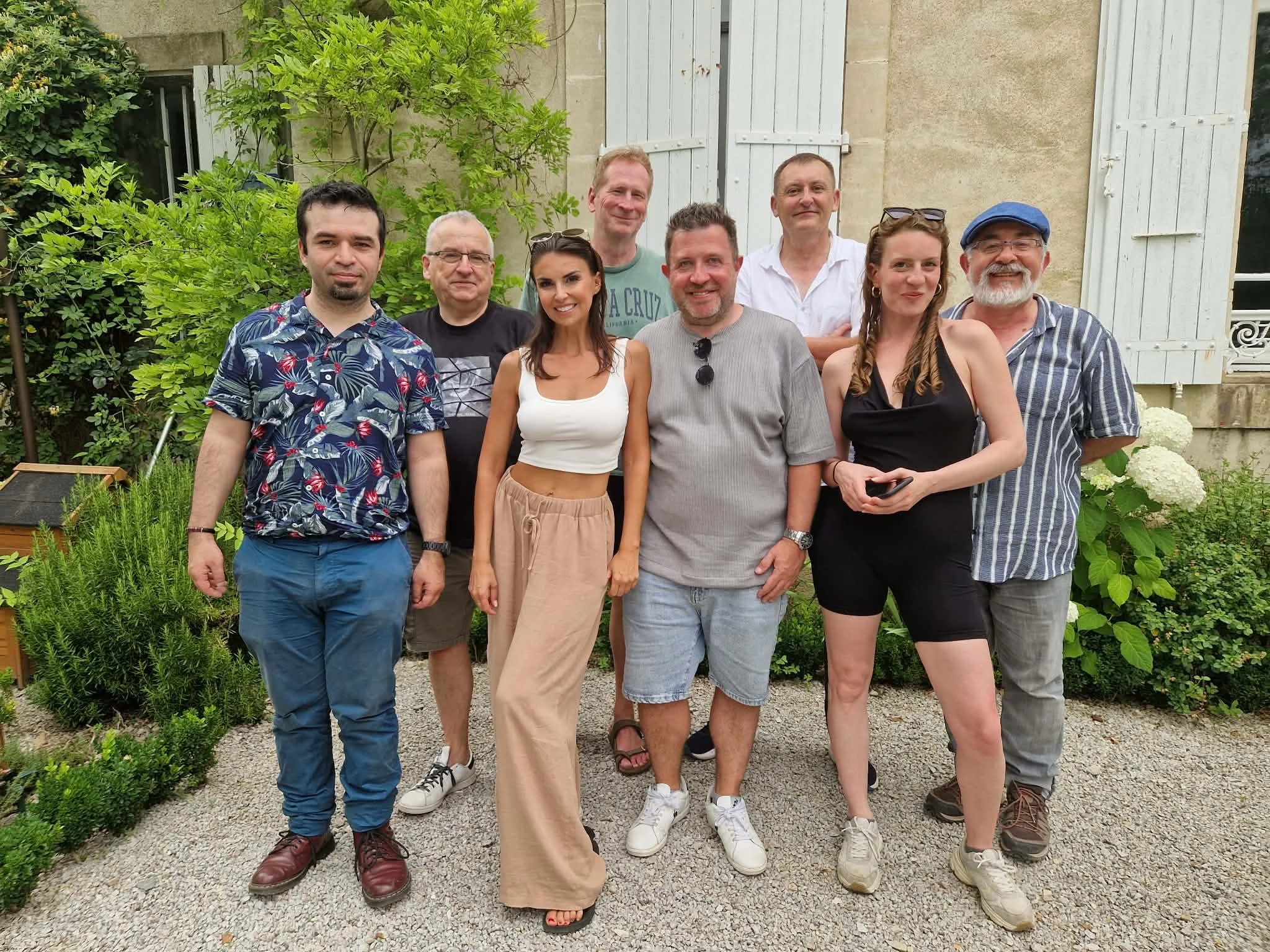 Groupe de neuf personnes posant devant une maison avec des volets en bois blanc, entourées de verdure et de fleurs, pendant une journée ensoleillée.