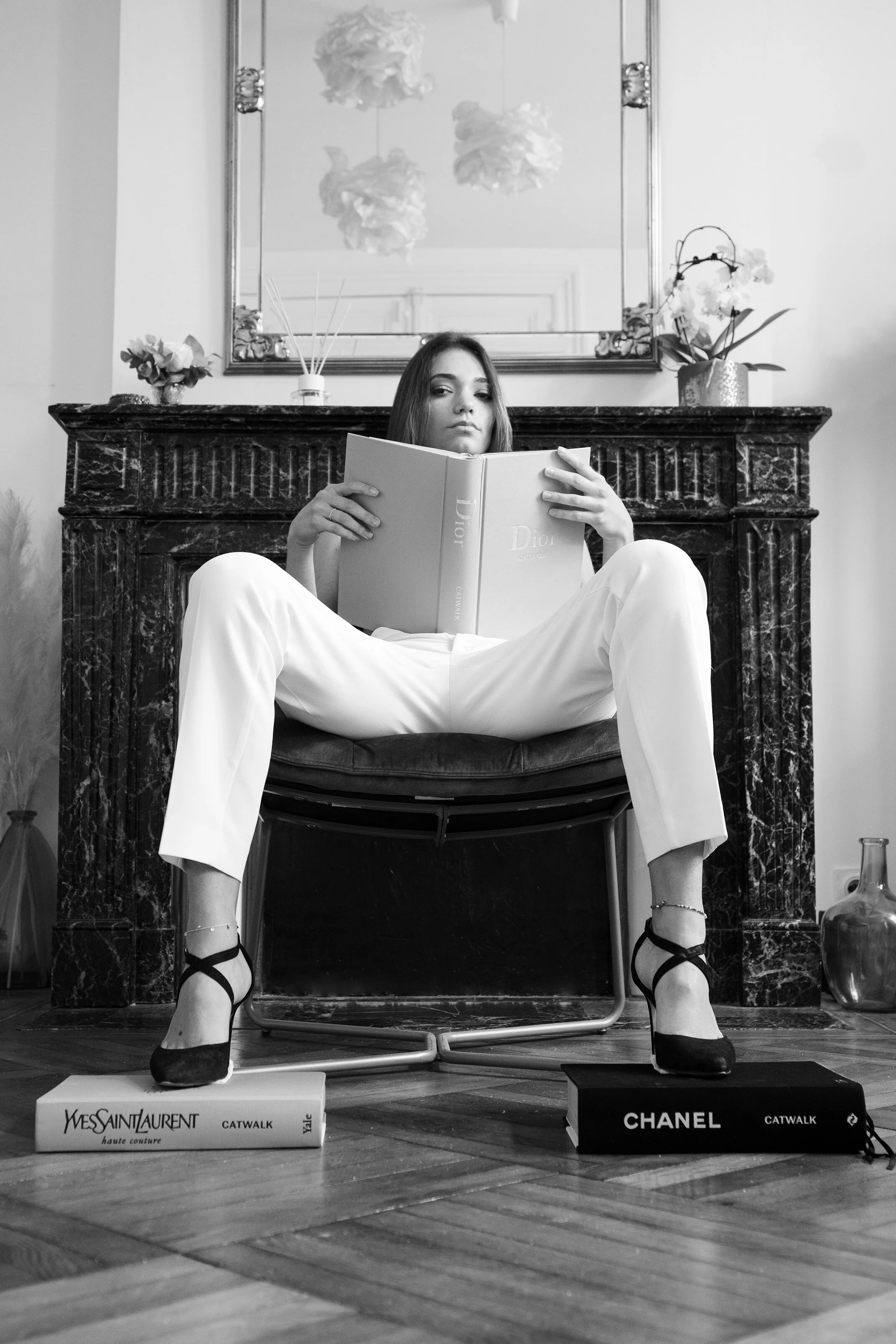 Une femme assise sur un fauteuil, portant des chaussures à talons noirs, lit un livre Dior. Deux grands livres, Yves Saint Laurent et Chanel, sont placés sous ses pieds.