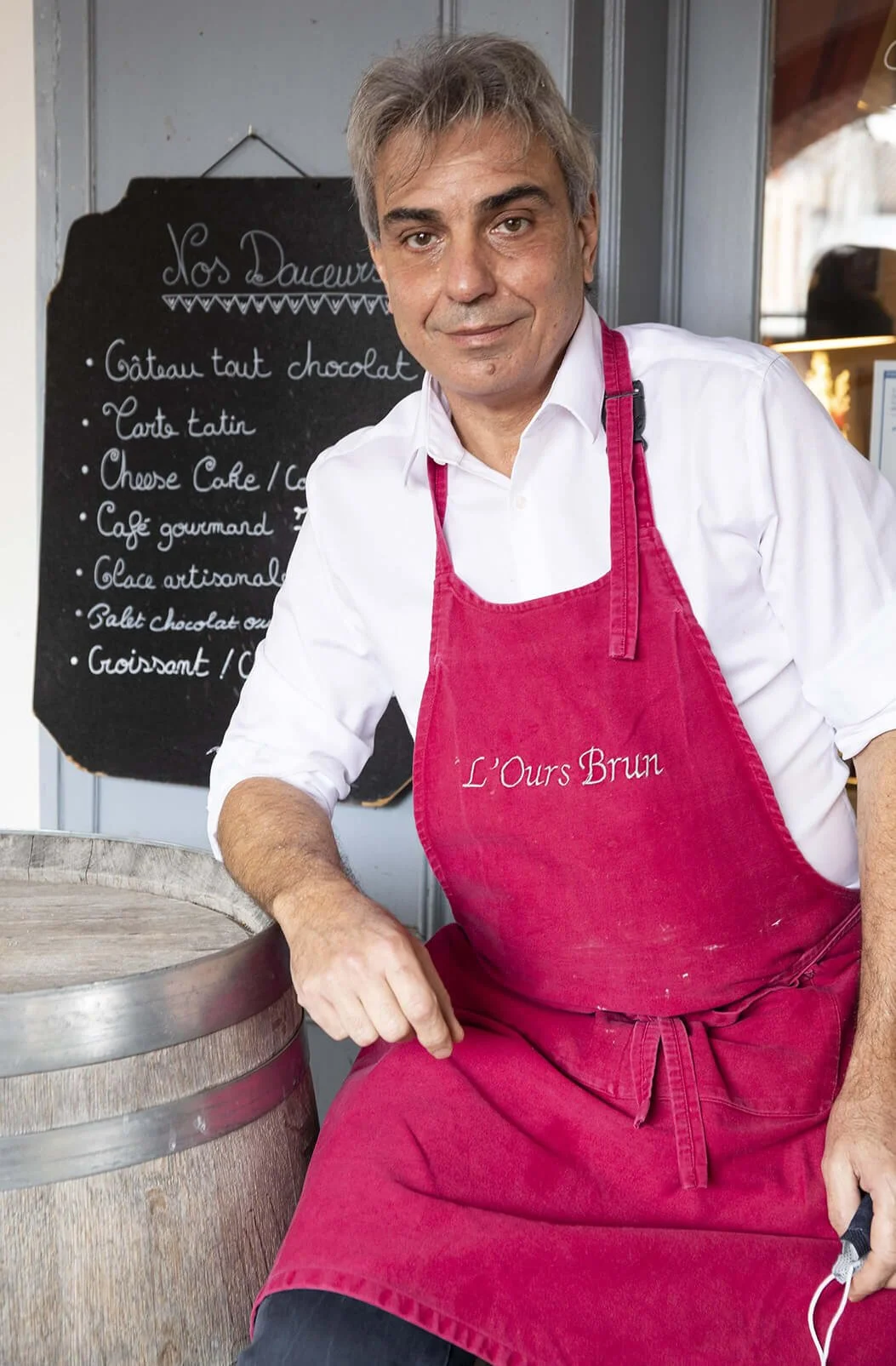 Un photographe professionnel immortalise un artisan pâtissier devant sa boutique.  Le portrait met en scène un homme en tablier rouge, soulignant le style photographique dans un cadre artisanal.  Le décor d’une pâtisserie accueille un artisan sourian