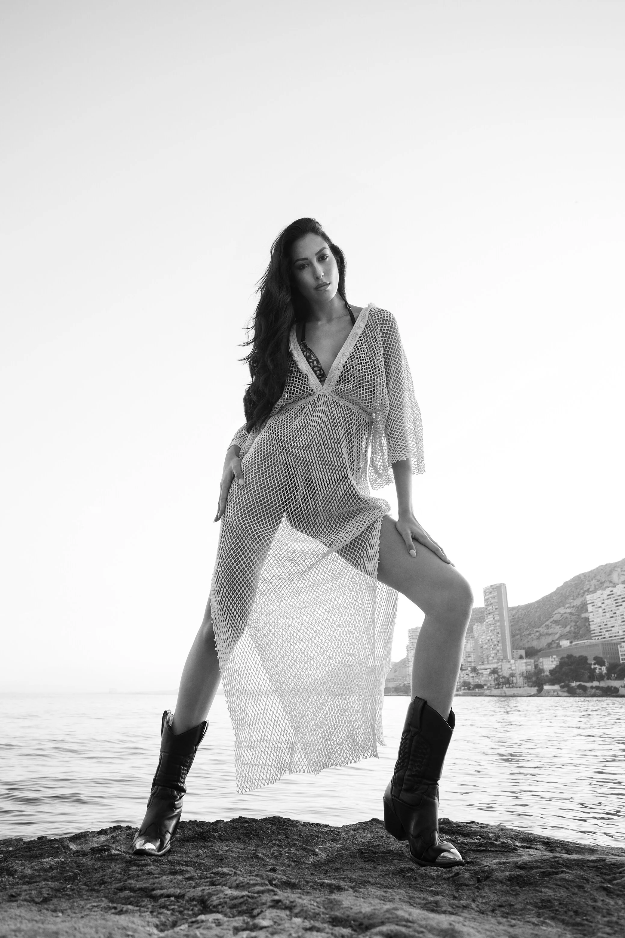 Une femme portant une robe à filet, des bottes noires, posant sur une surface rocheuse près de l'eau, avec une ville et des montagnes en arrière-plan, en noir et blanc.