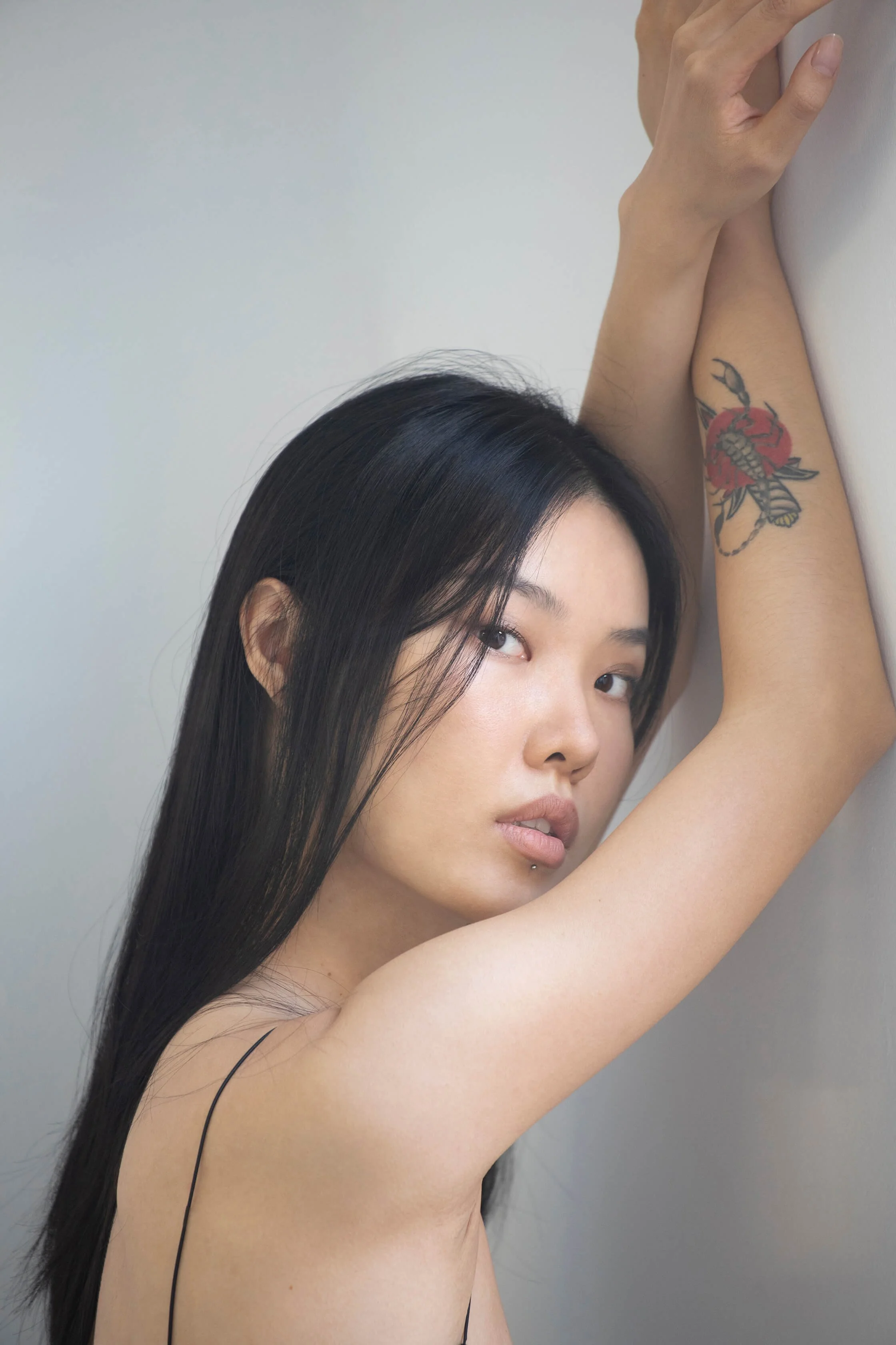 Jeune femme aux cheveux longs et noirs, avec tatouage de rose sur le bras, pose contre un mur, regardant vers l'objectif.