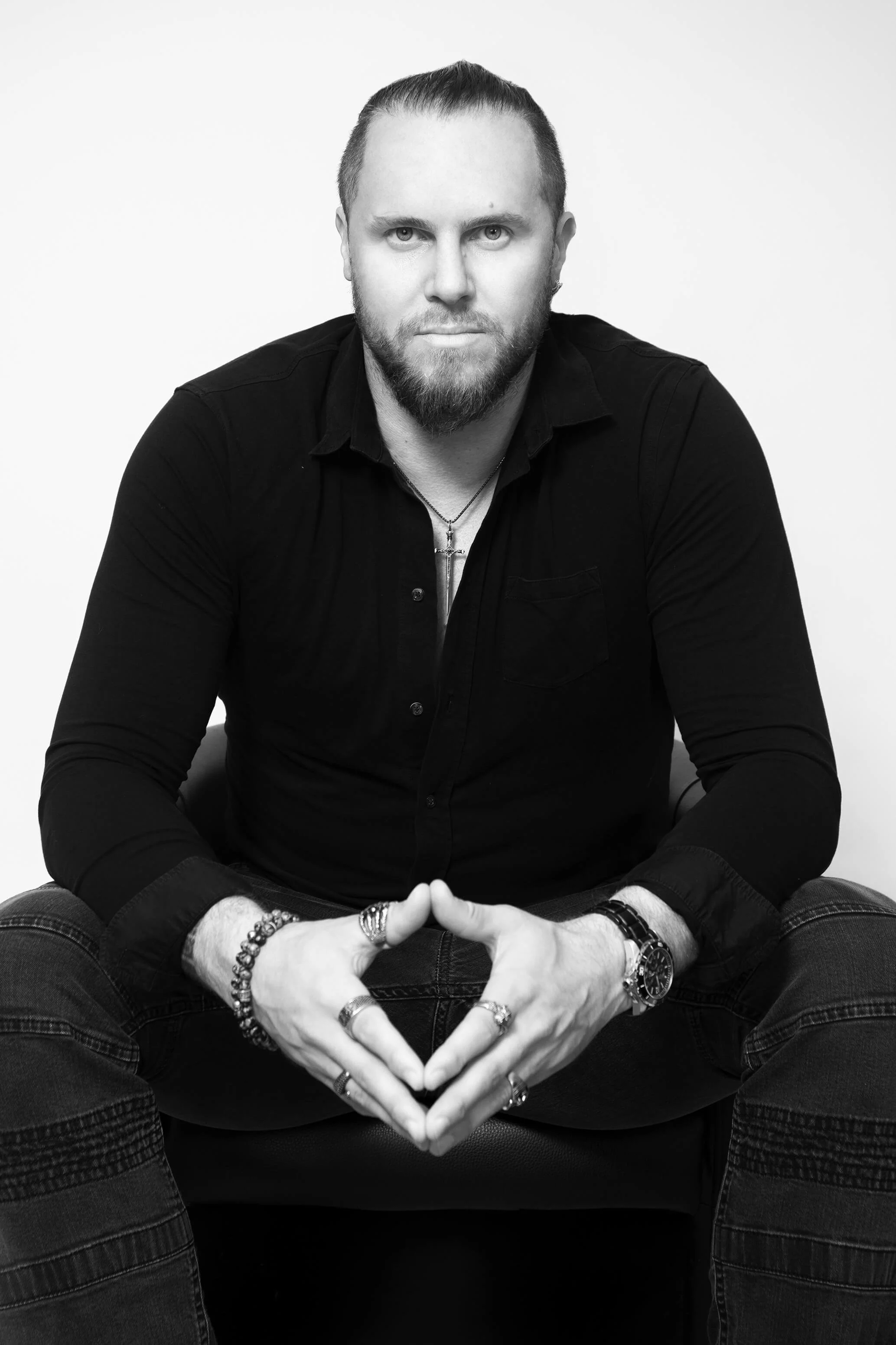 Homme avec barbe, portant des bracelets et une montre, assis en position de méditation, formant un cœur avec ses mains, habillé en noir, fond blanc.