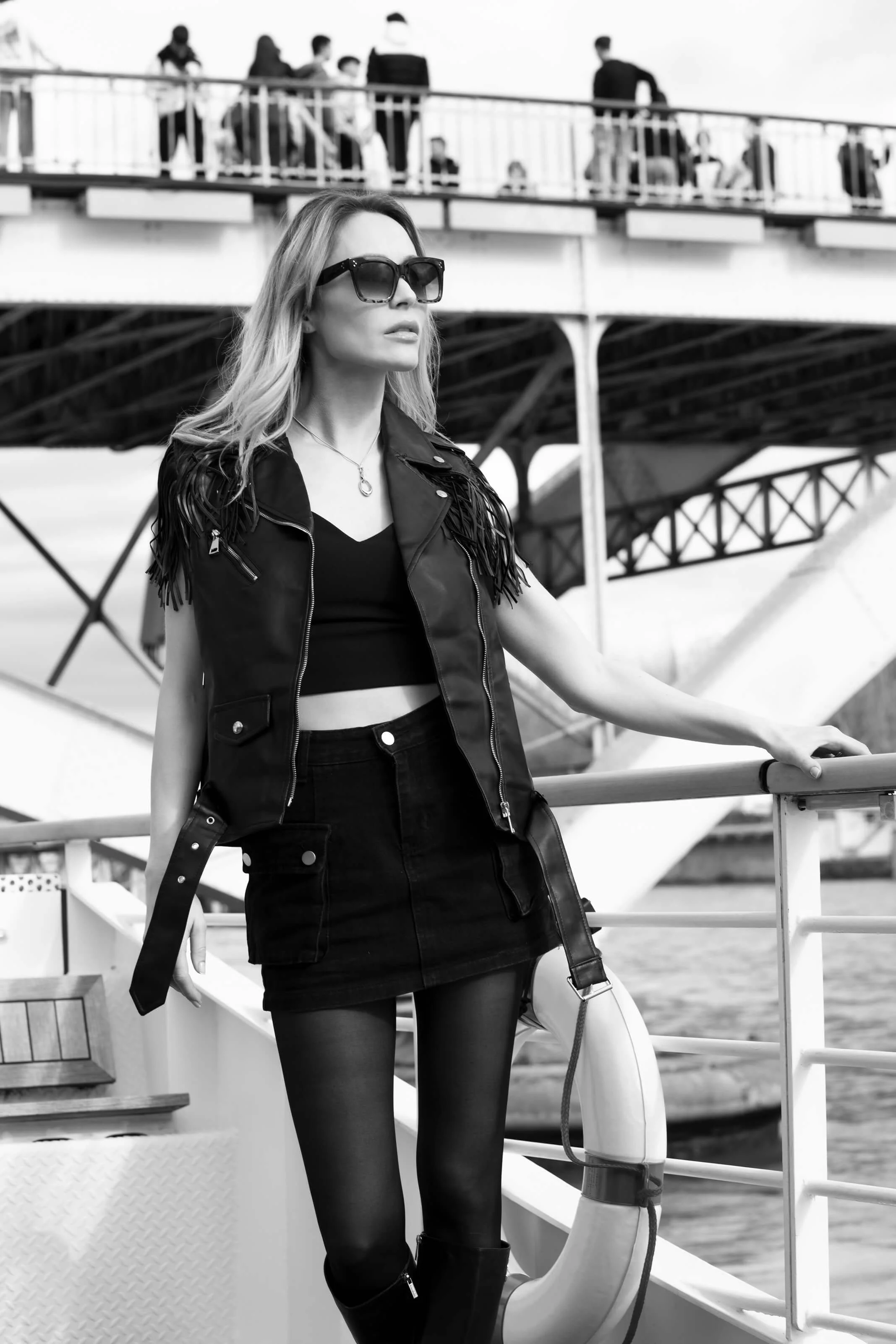 Une femme portant des vêtements noirs, des lunettes de soleil et un sac à main, en costume de mode, se tenant sur le pont d'un bateau, avec une structure métallique et un groupe de personnes en arrière-plan.