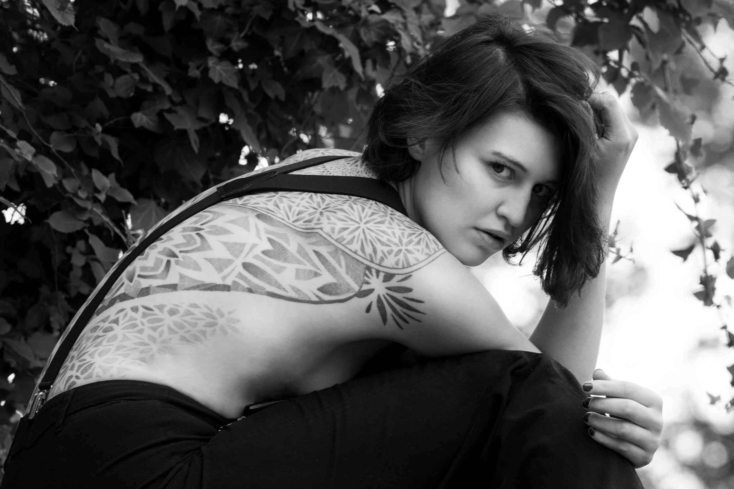 Une femme avec un tatouage complexe sur l'épaule et le bras, assise à l'extérieur près de feuillage dense. Elle a les cheveux courts, foncés, et regarde vers l'objectif avec une expression sérieuse.