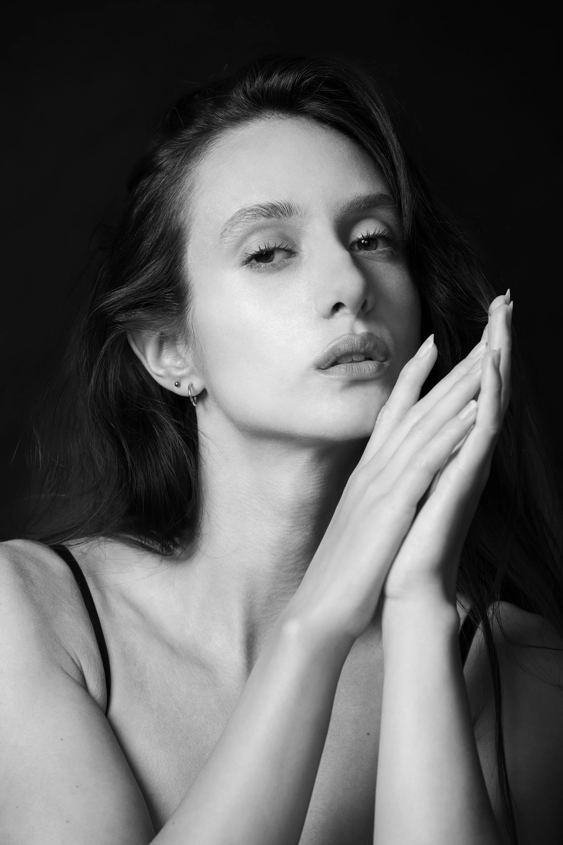 Portrait en noir et blanc d'une femme avec des longs cheveux, portant des boucles d'oreilles, yeux mi-clos, et mains jointes près du visage.
