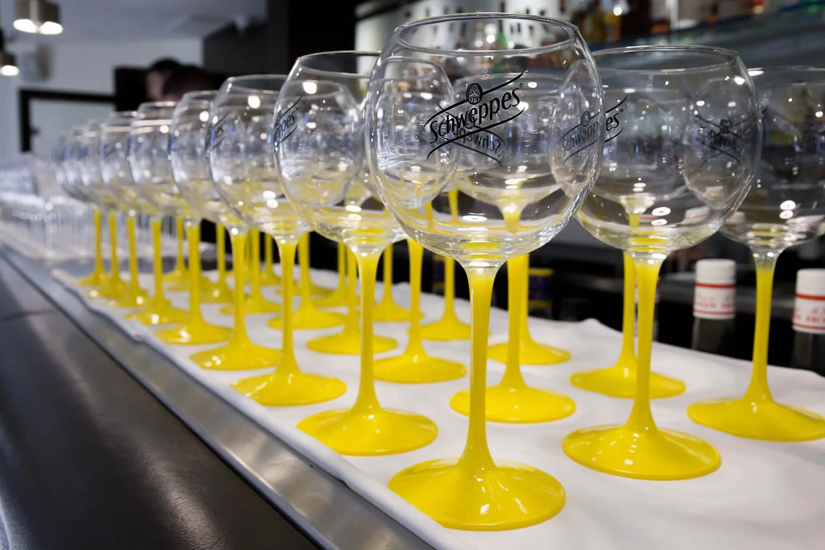 Des verres à pied jaunes de Schweppes, alignés avec élégance, ornent le bar pour un événement festif.  Le style photographique de François Fauré met en lumière ces verres modernes, créant une ambiance événementielle raffinée.  Sur un bar contemporain