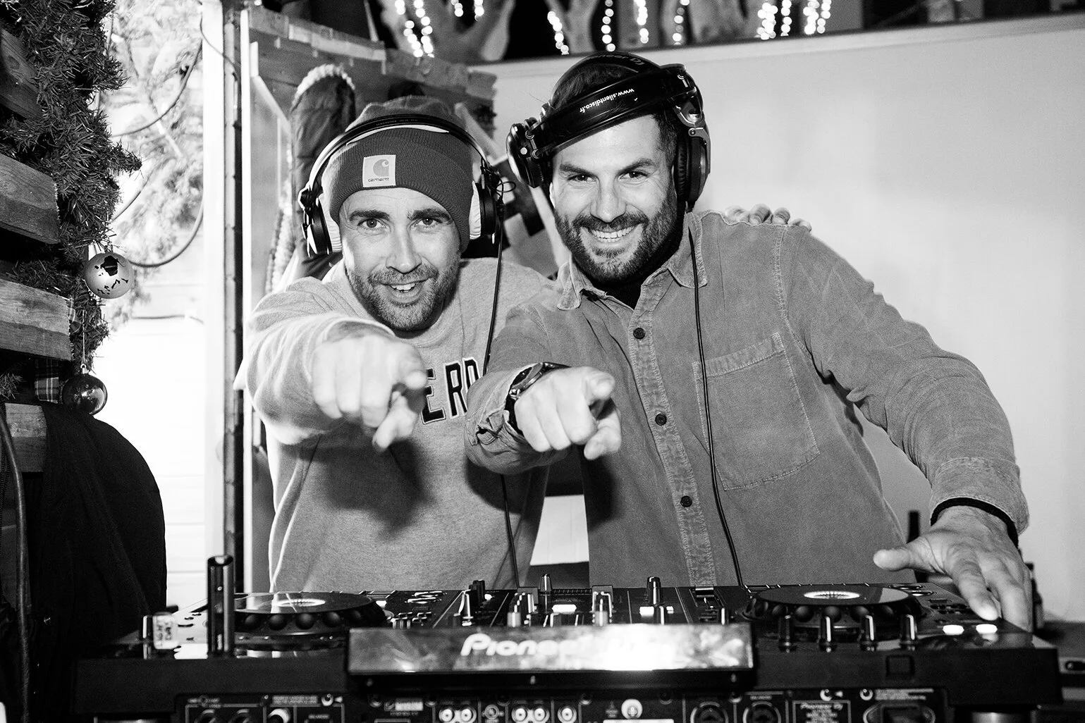 Deux DJs souriants, plongés dans l’ambiance festive, pointent vers l’objectif.  L’image en noir et blanc immortalise leur performance, capturant l’énergie de la soirée musicale.  Le photographe professionnel immortalise l’événement, mettant en valeur