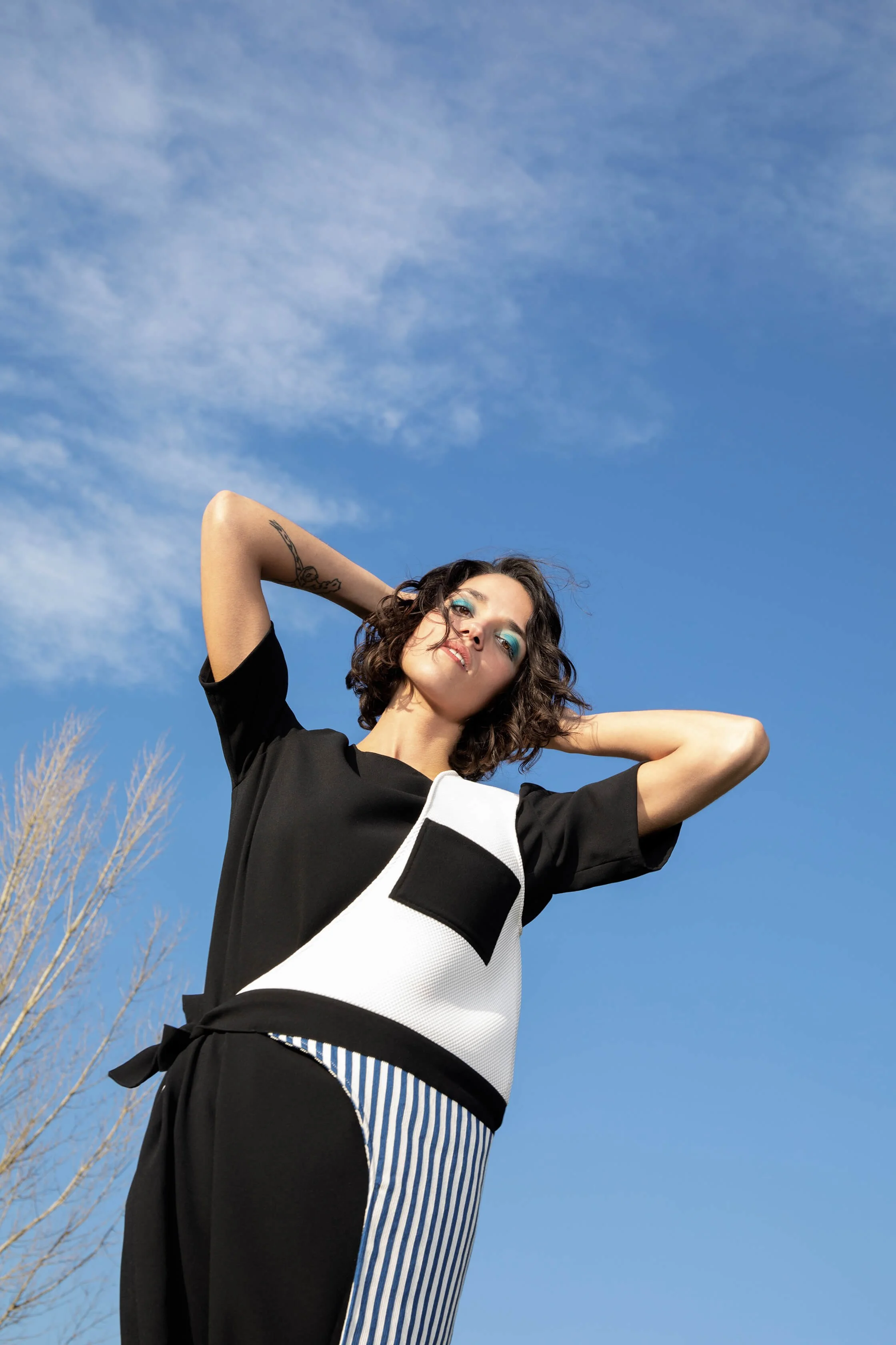 Femme posant en tenue moderne avec un haut noir et un pantalon rayé sous un ciel bleu.
