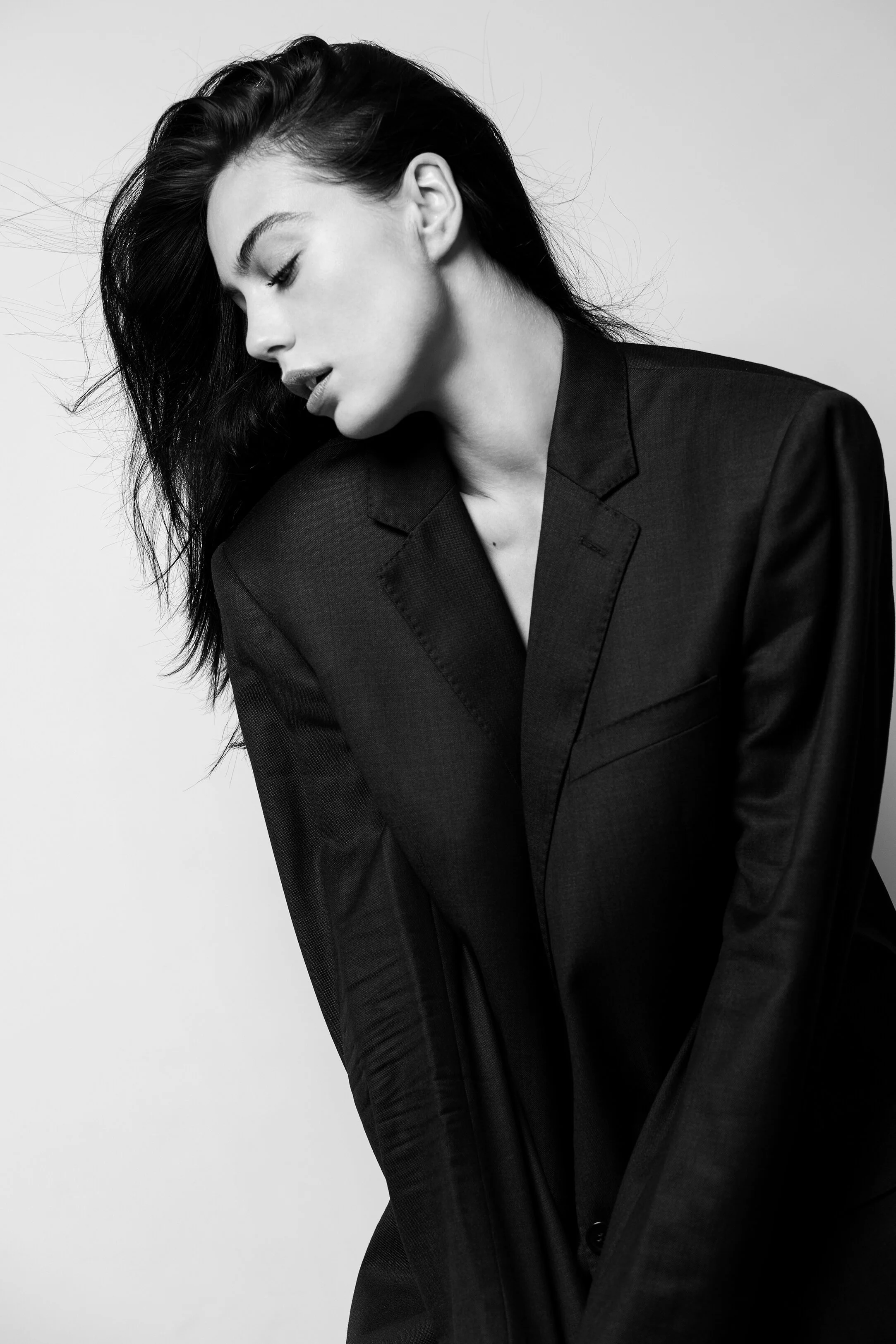 Une femme en noir et blanc portant un blazer noir, regardant vers le sol avec les yeux fermés, cheveux noirs longs et coiffés de manière ondulée.
