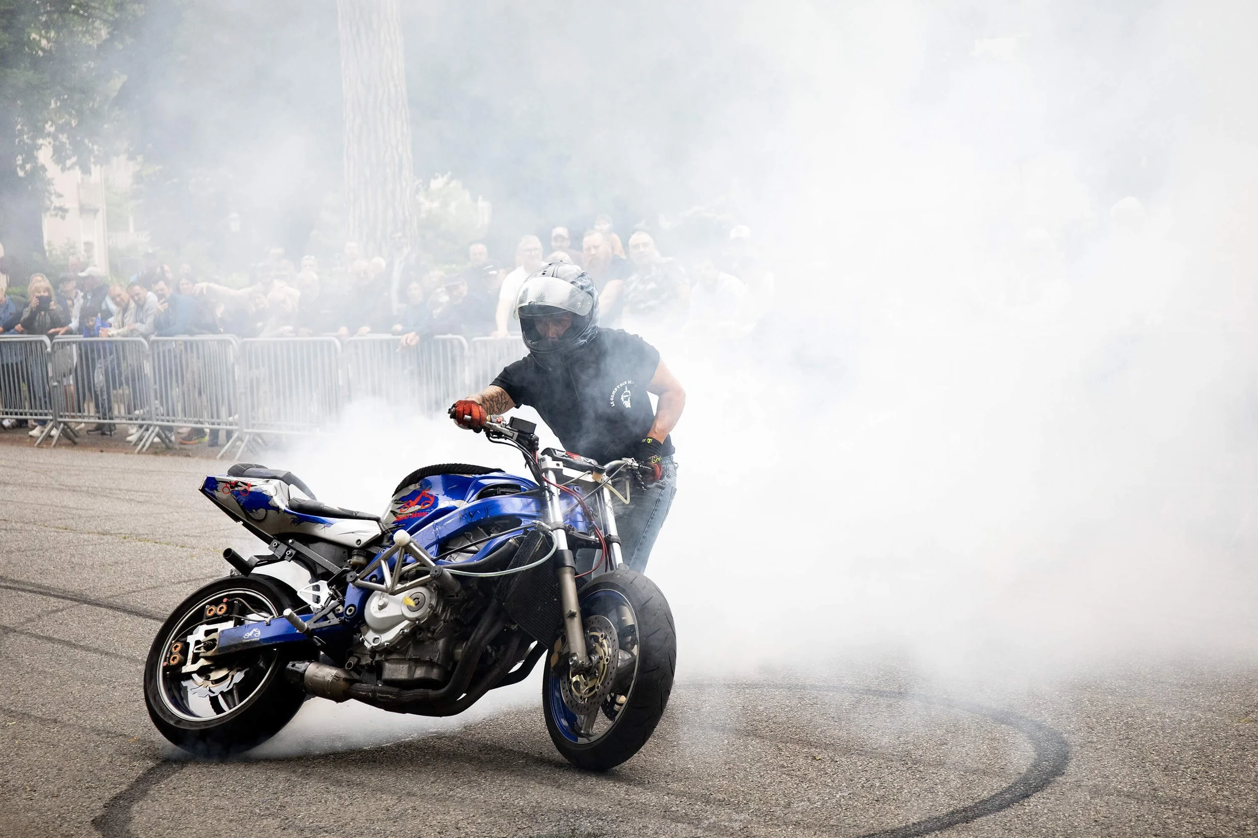 Un pilote de moto exécute des figures acrobatiques dans un nuage de fumée lors d’un événement. Le public est captivé par les motos en action. Un motard effectue une roue arrière sur une moto bleue, entouré de spectateurs. La performance de stunt crée