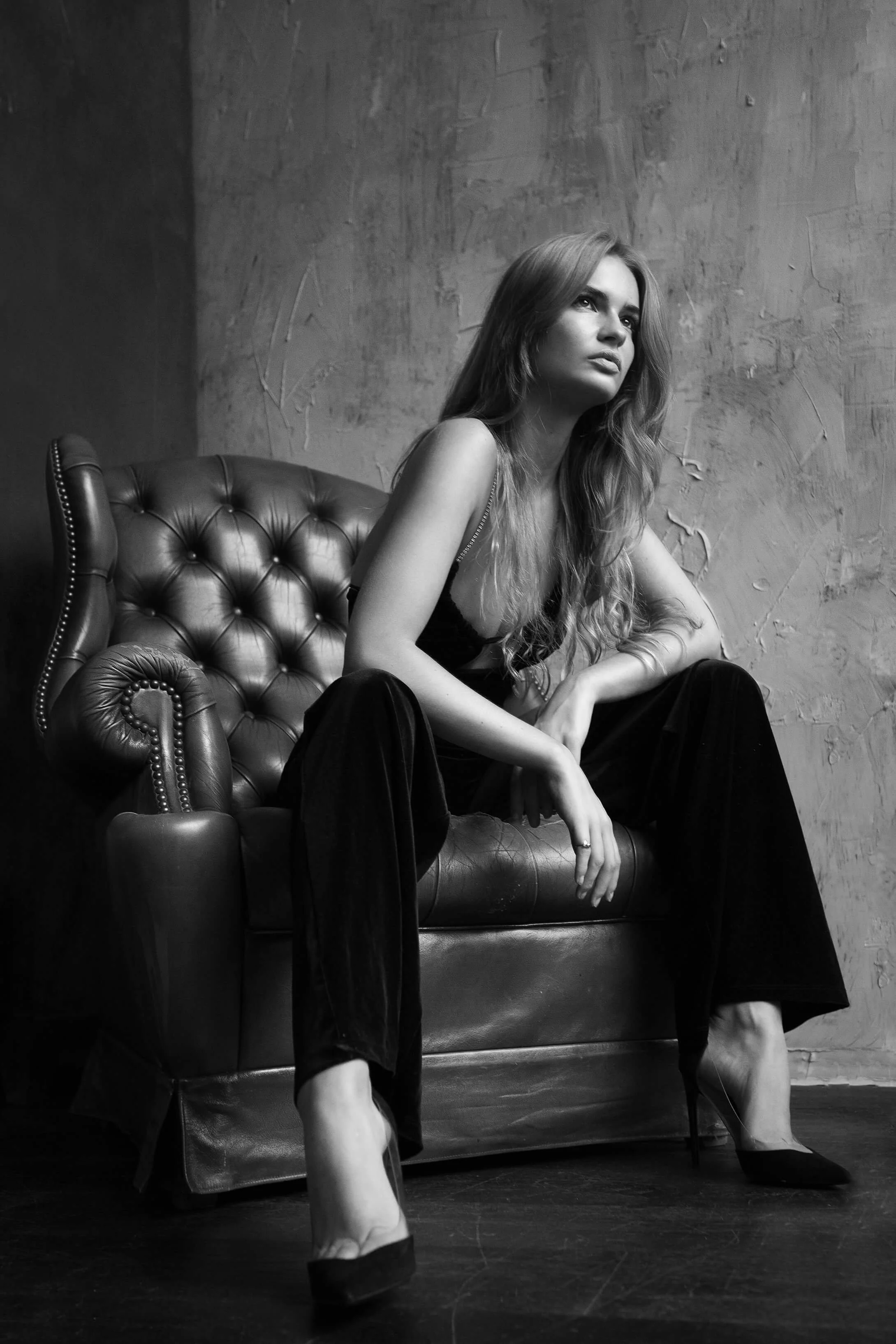 Une femme assise sur un canapé en cuir, en costume noir, dans une pièce avec un mur gris textured, en noir et blanc.