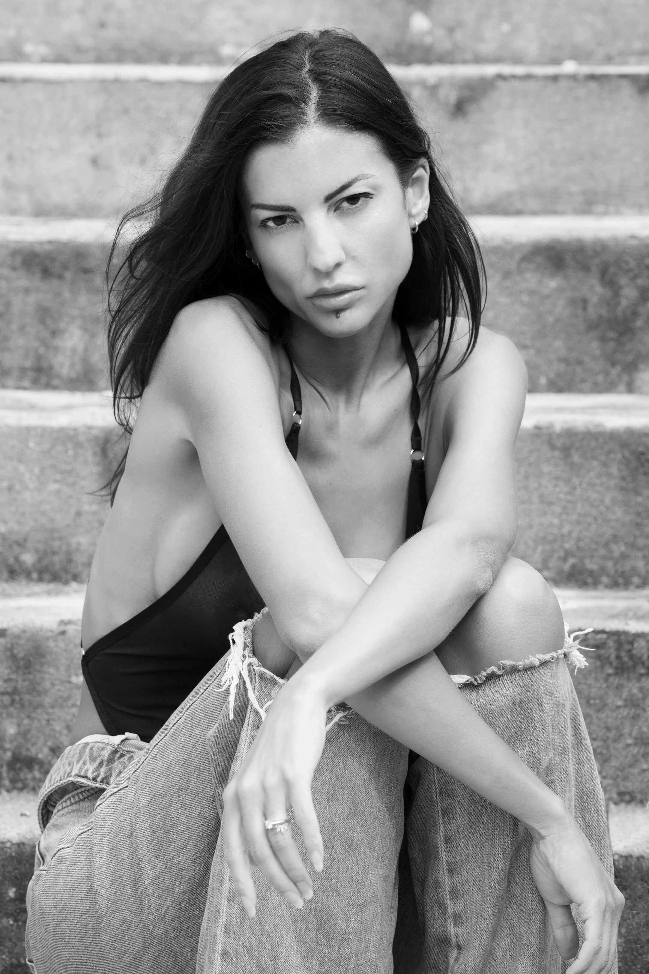 Une femme aux cheveux longs et foncés, assise sur des escaliers, portant un haut noir et un jean déchiré, avec une expression sérieuse.