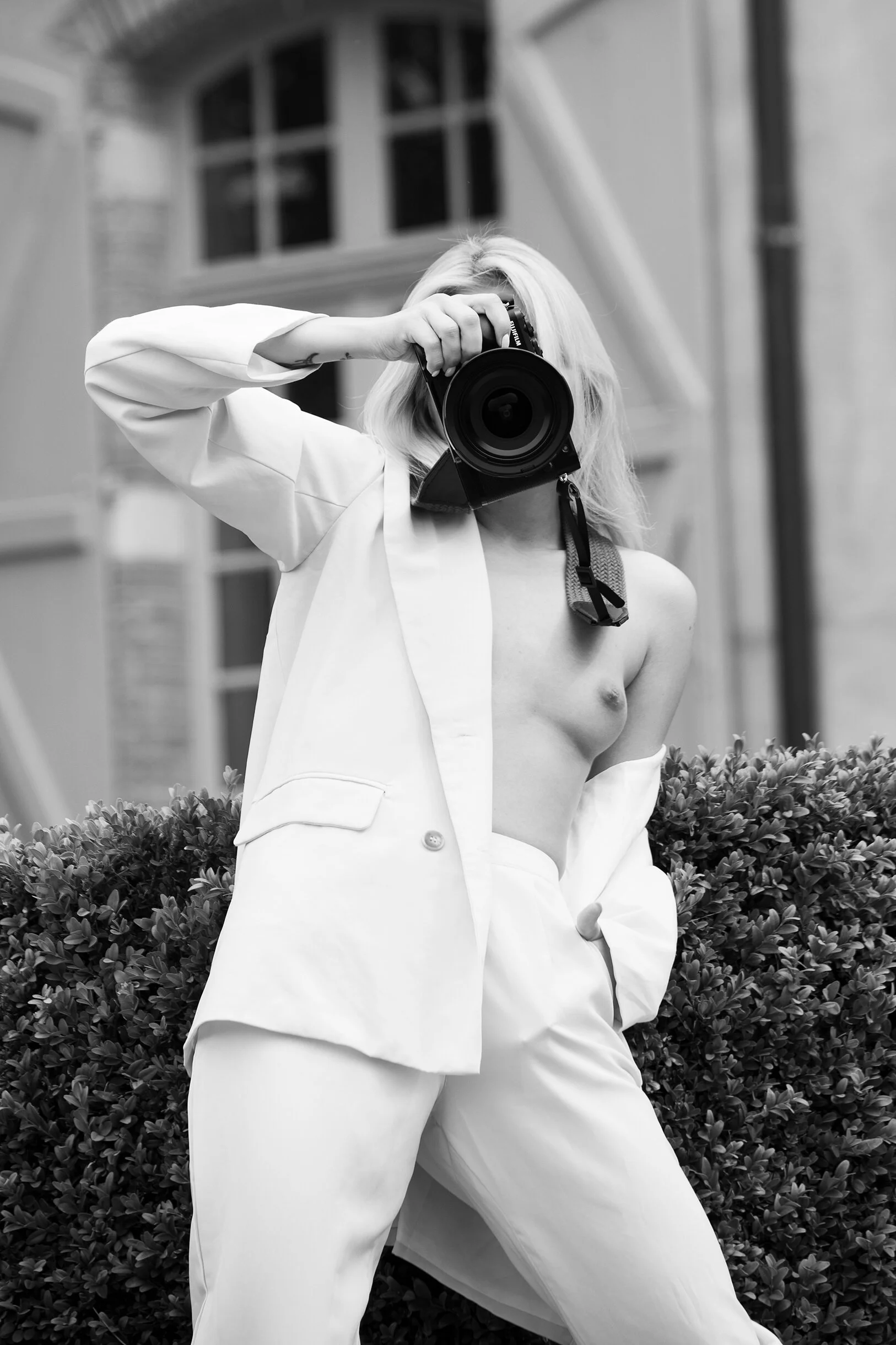 Une personne portant un costume blanc non porté, tenant un appareil photo, en extérieur, en noir et blanc.