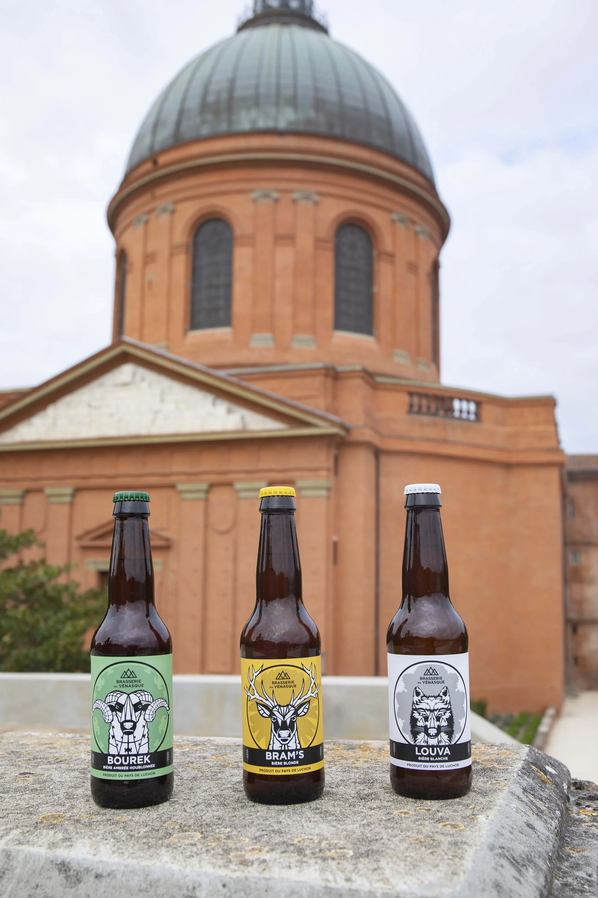 Des bouteilles de bière artisanale se dressent devant un bâtiment historique, créant une composition photographique captivante.  Le fond architectural met en valeur les bières, tandis que les étiquettes colorées contrastent avec l’édifice ancien.  Un
