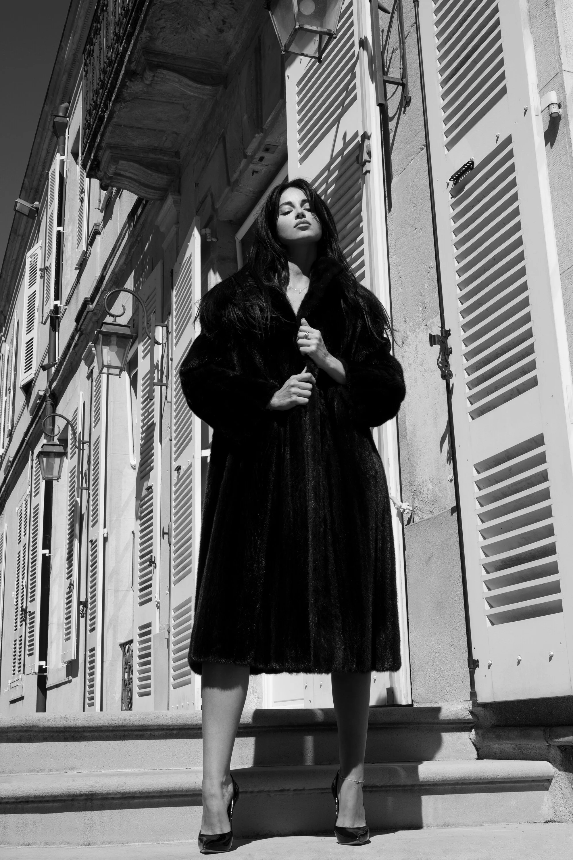 Une femme élégante en manteau noir long pose devant une façade de bâtiment avec des volets blancs, prise en noir et blanc.