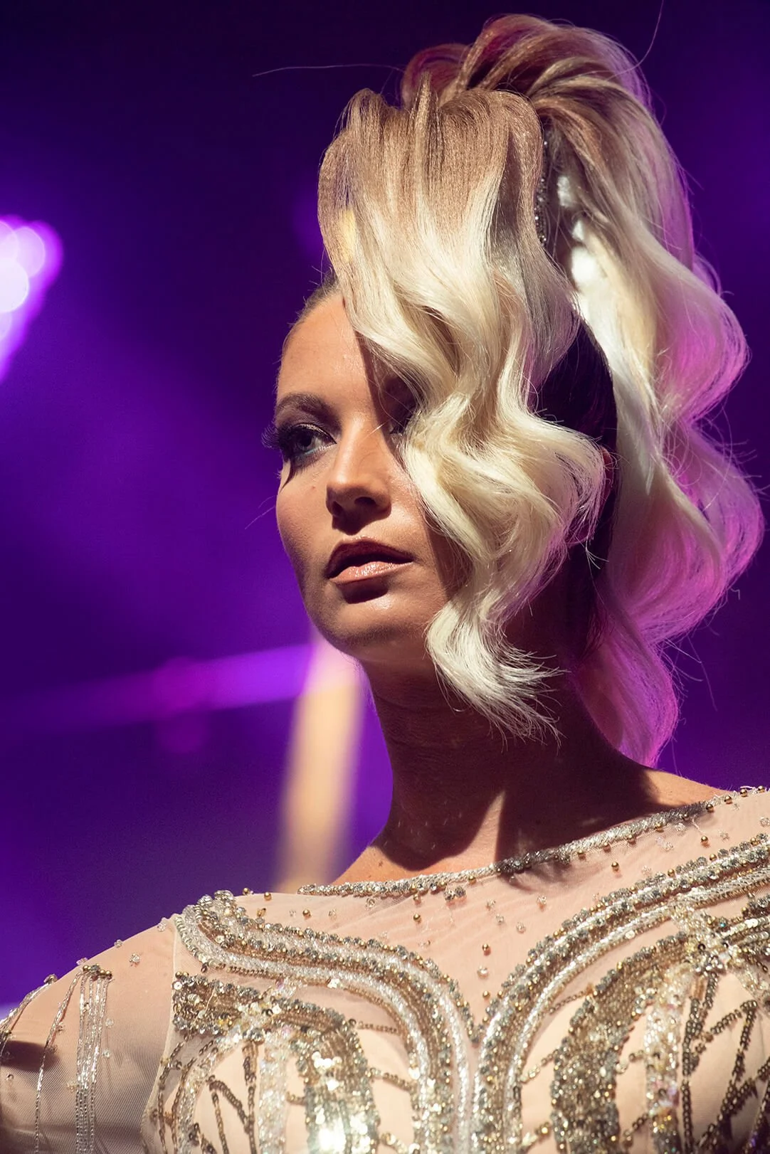 Portrait d'une modèle avec une coiffure artistique, mettant en valeur des couleurs de cheveux blonds et un décor lumineux.
Vue rapprochée d'une coiffure élégante, accentuée par des lumières violettes et des détails scintillants.
Modèle présentant une
