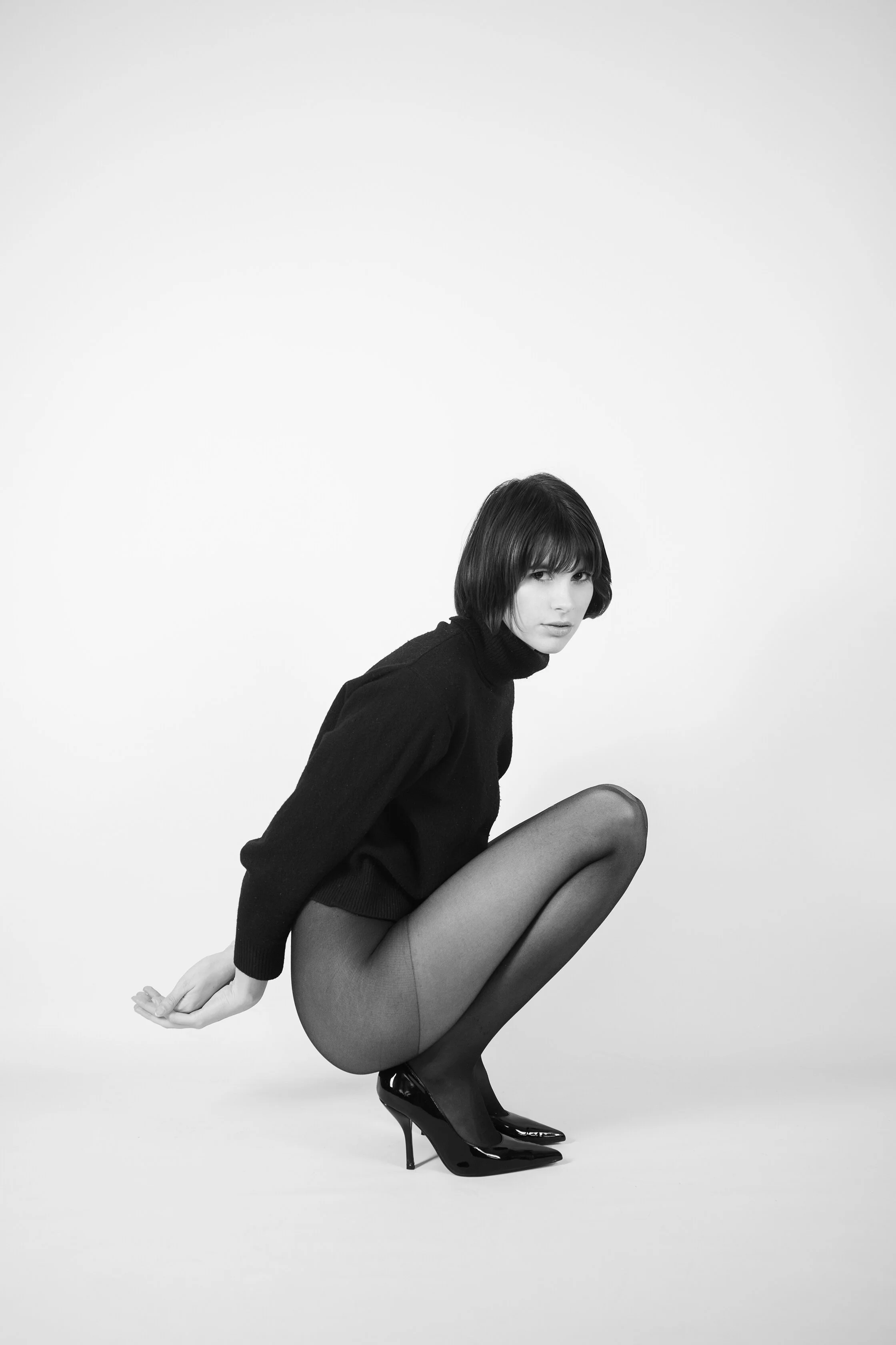 Une femme en noir et blanc portant un pull noir, des collants et des talons noirs, en position accroupie sur un fond blanc.