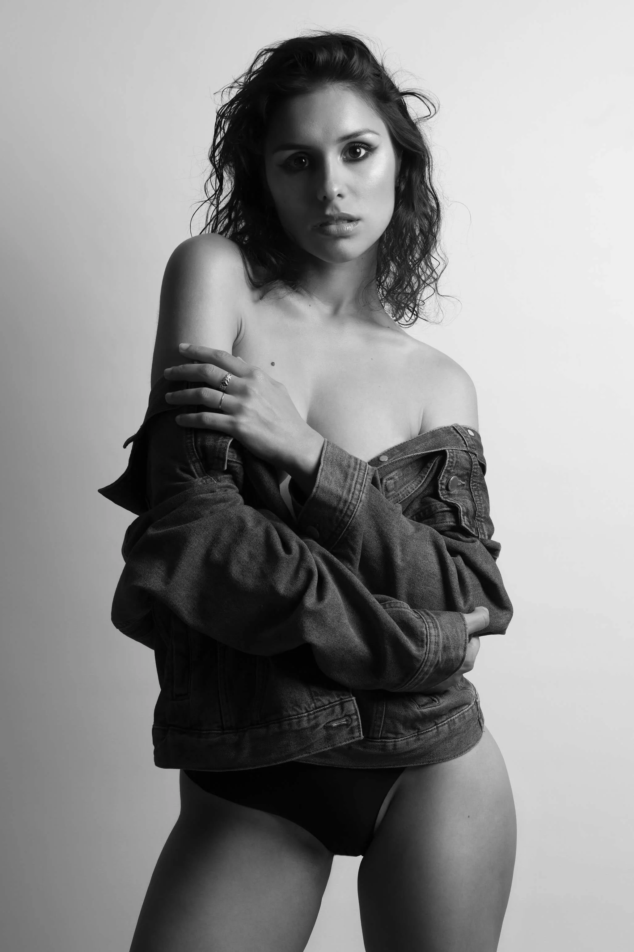 Une femme pose en lingerie noire avec un jean porté en veste, dans un fond gris. La photo est en noir et blanc.