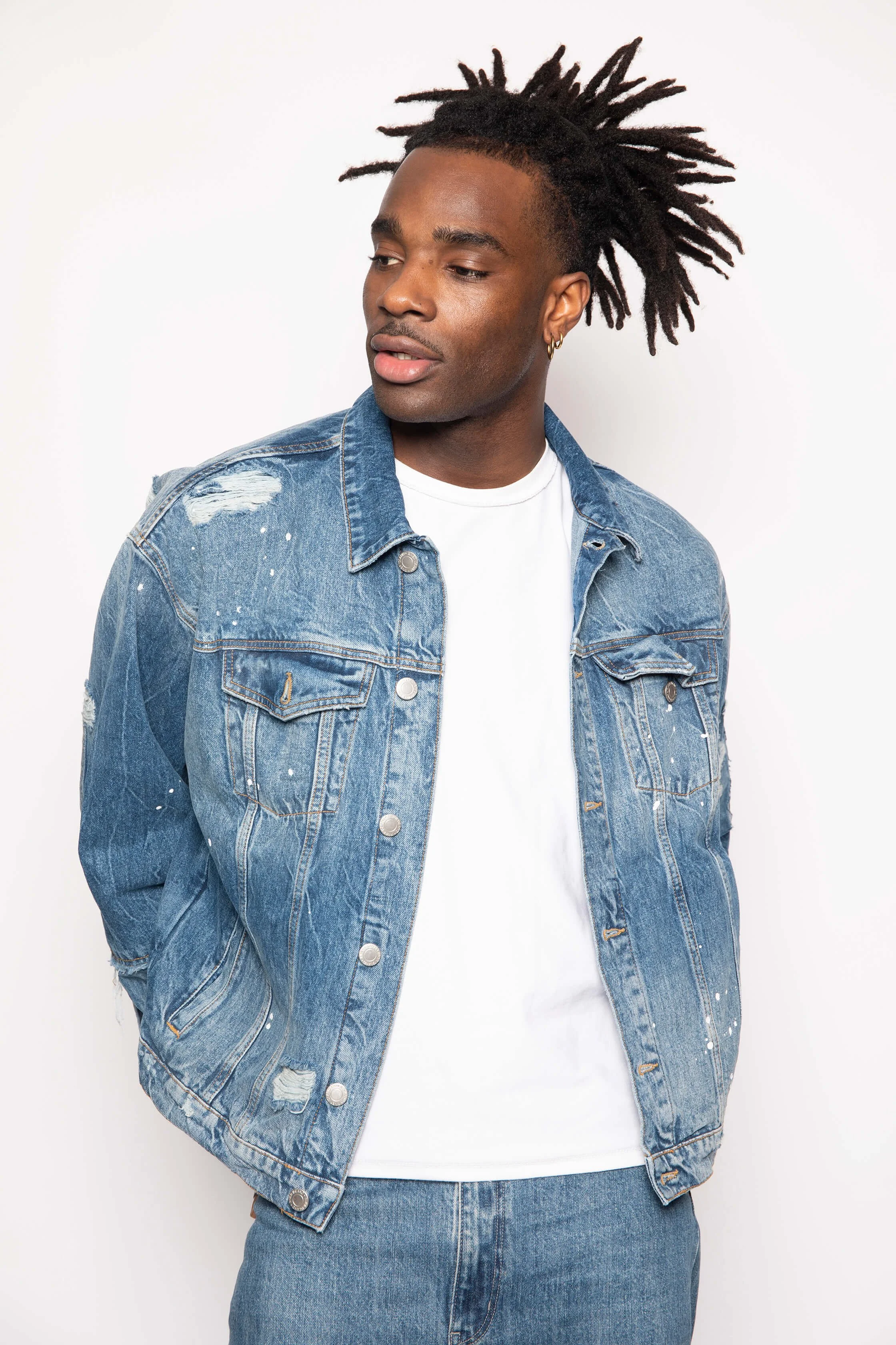 Jeune homme portant une veste en jean et un t-shirt blanc, regardant vers la gauche, contre un fond blanc.