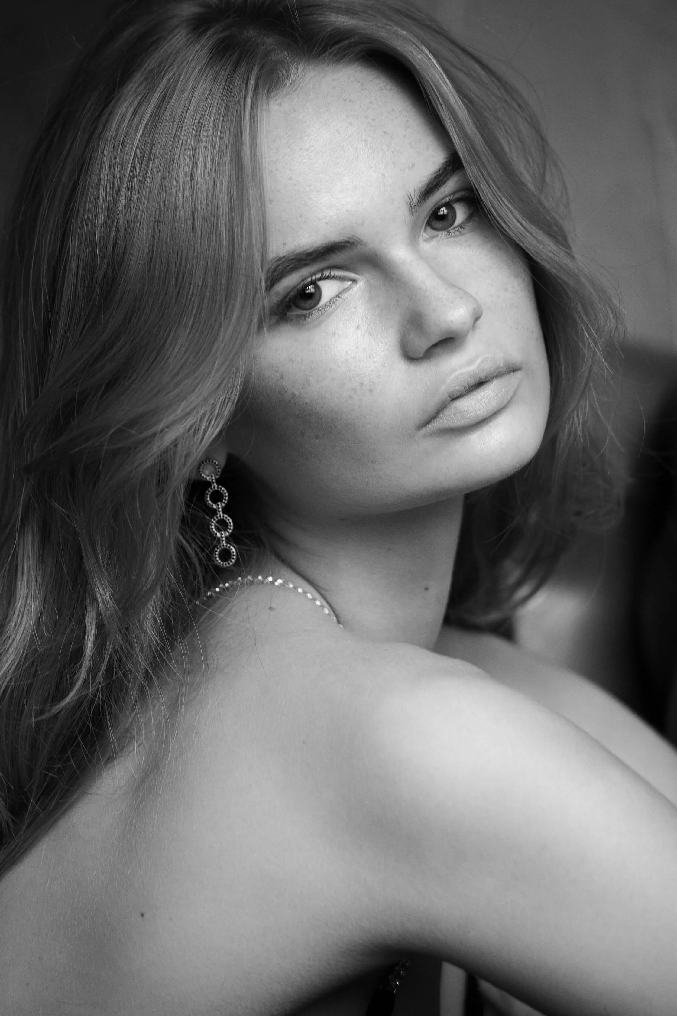 Portrait en noir et blanc d'une jeune femme avec des cheveux ondulés, portant une boucle d'oreille et un collier, regardant vers l'objectif.