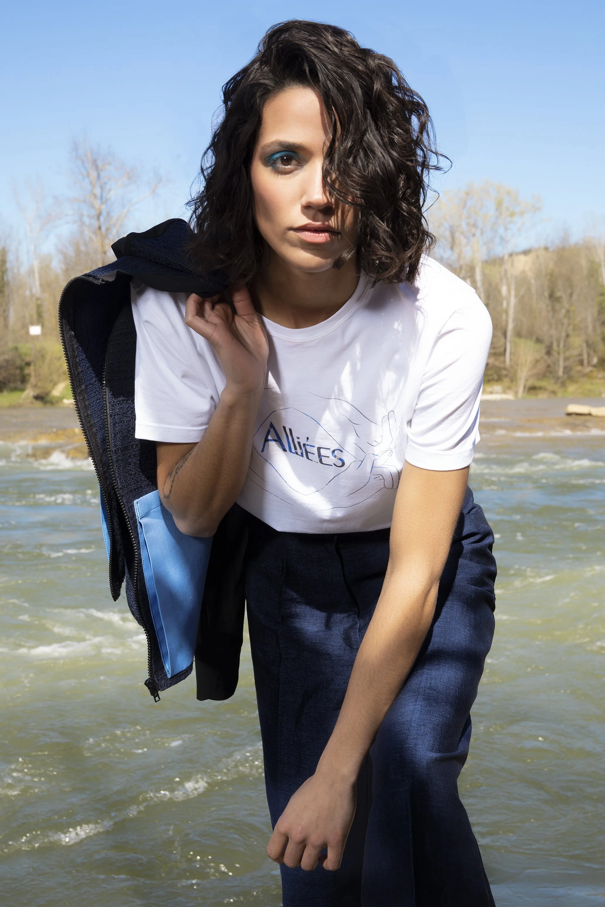 Femme posant près d'une rivière, vêtue d'un t-shirt blanc et d'un pantalon bleu, avec une veste sur l'épaule