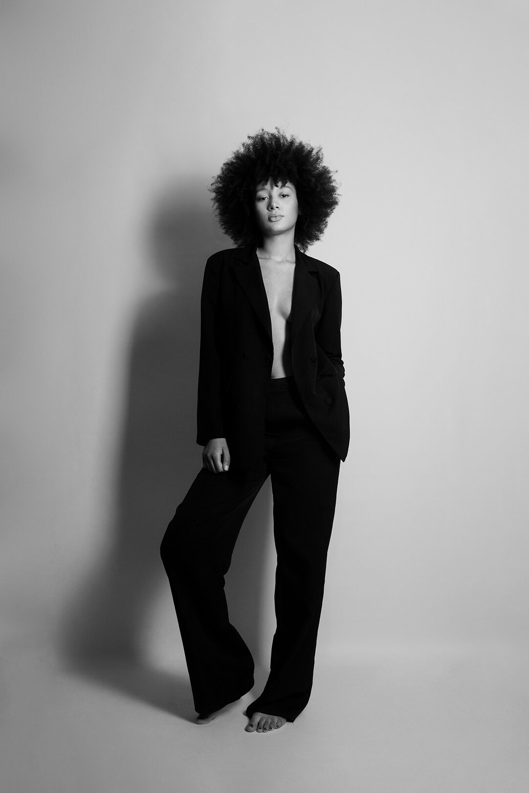 Une femme aux cheveux afro porte un costume noir sans chemise, debout sur un fond neutre, dans une photographie en noir et blanc.