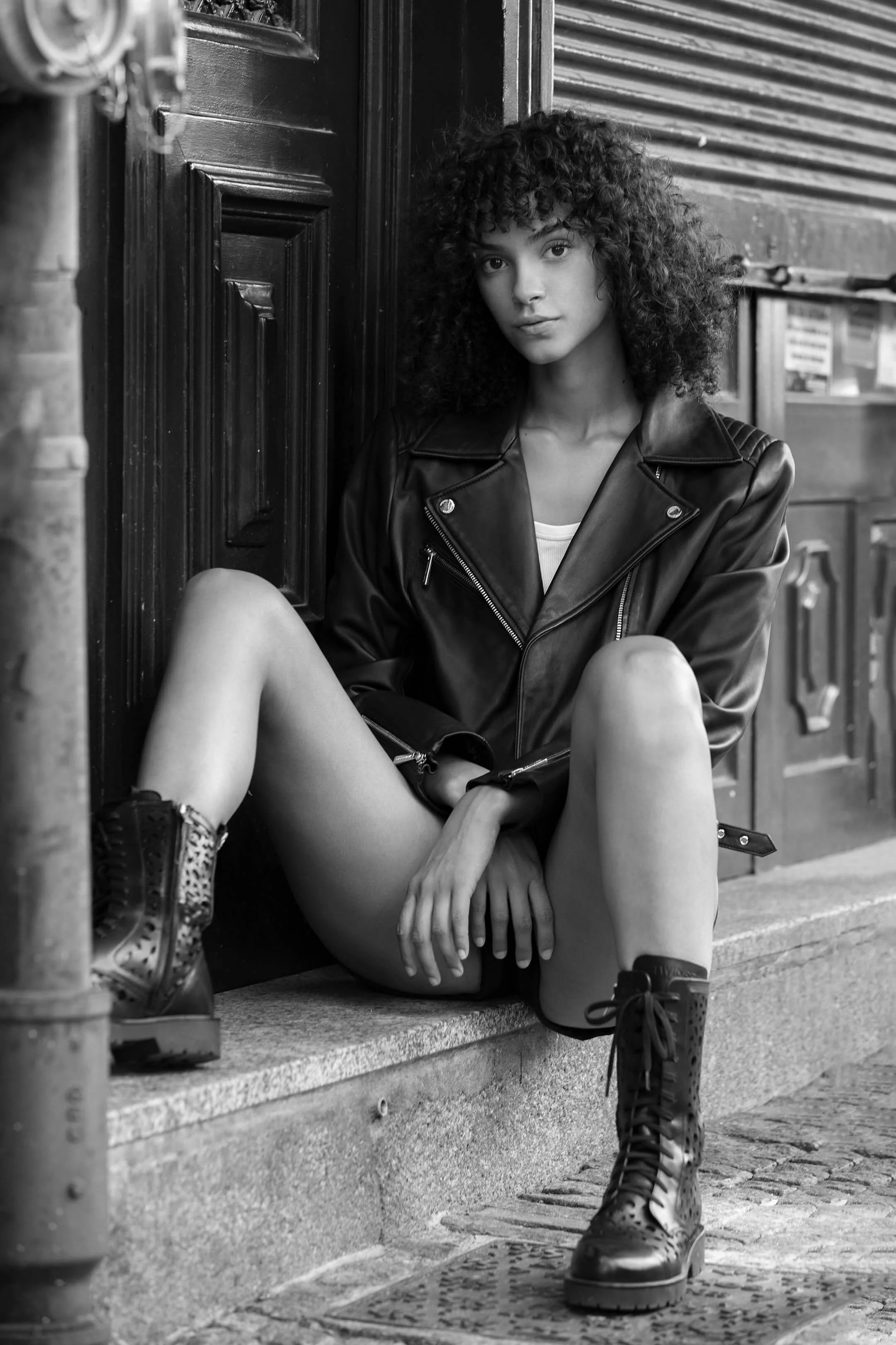 Une jeune femme avec des cheveux bouclés assise par terre en noir et blanc, portant une veste en cuir, des bottes à lacets, dans une rue urbaine