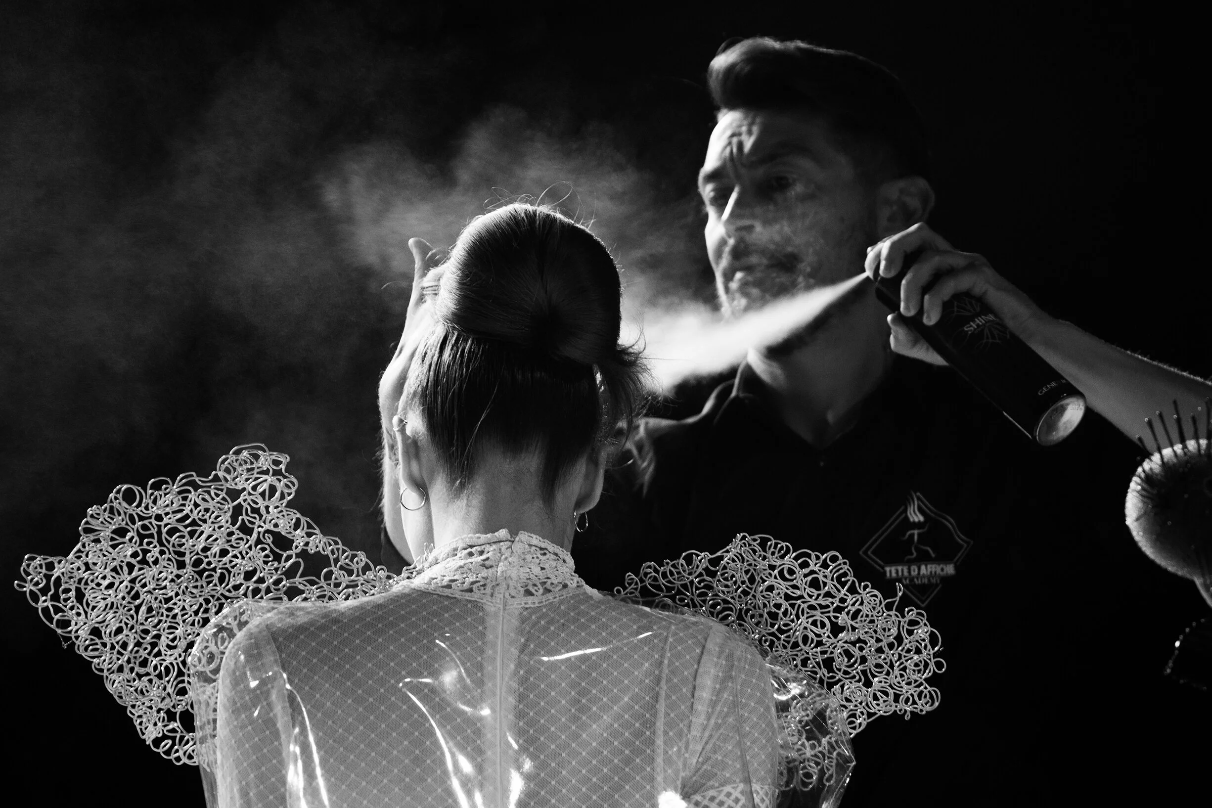 Un coiffeur applique un spray sur une coiffure créative, illustrant l'art de la coiffure dans un décor stylisé.