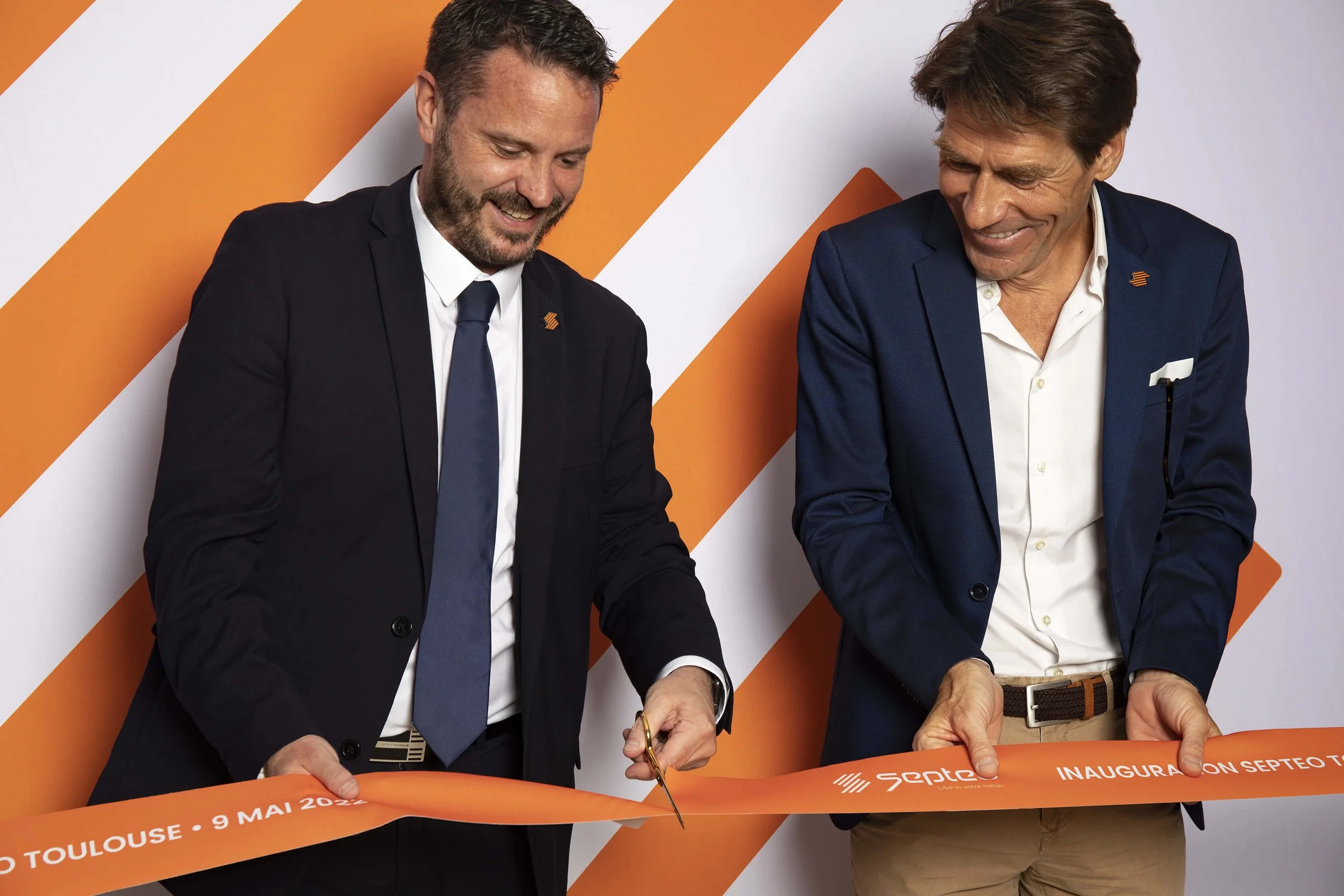 Deux hommes coupent un ruban lors d’une cérémonie d’inauguration à Toulouse, devant un fond orange. Le style photographique de François Fauré se distingue par ses couleurs vives, capturant parfaitement l’ambiance festive de l’événement.  L’image met 