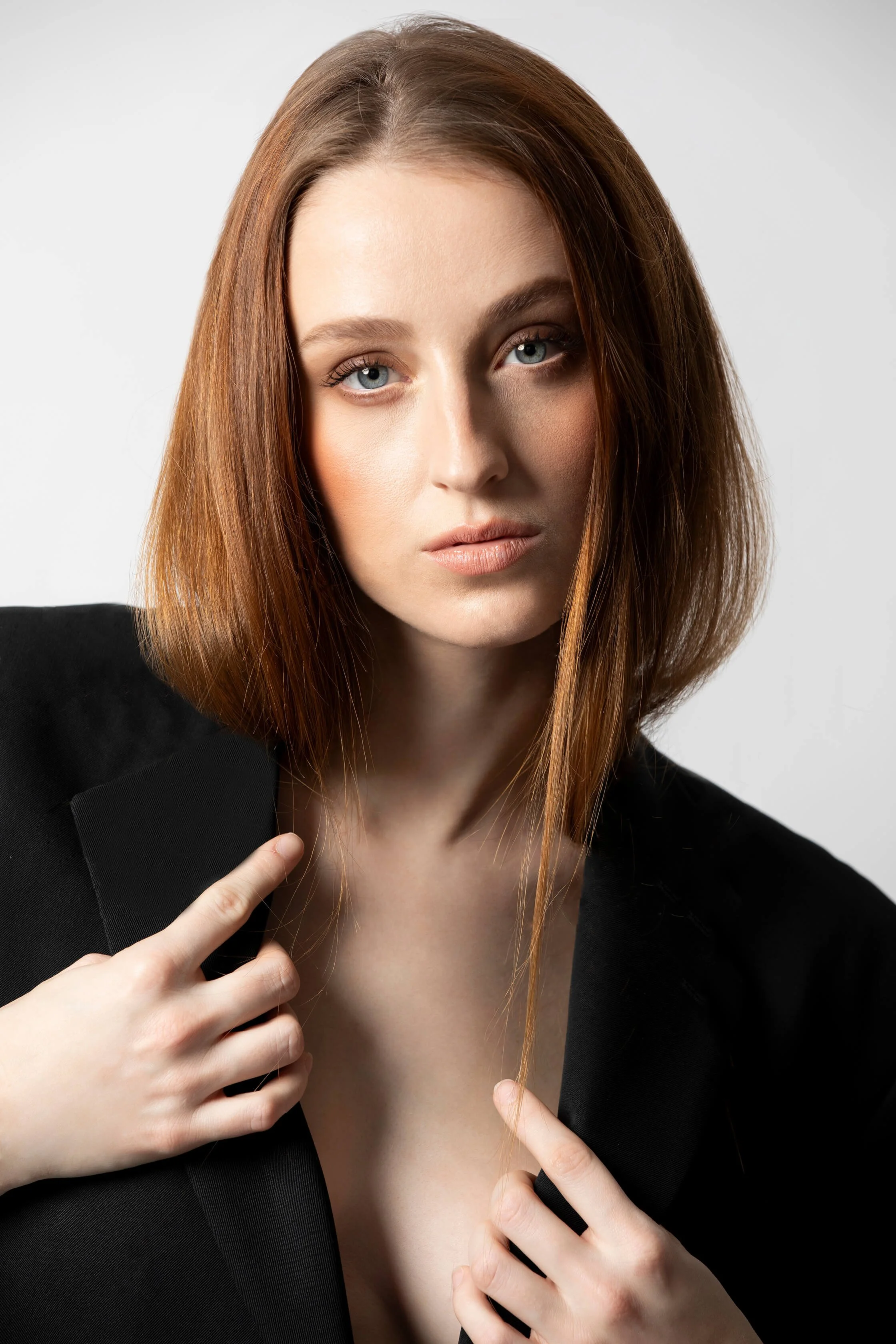 Femme aux cheveux roux, visage sérieux, portant un blazer noir, pointant le col de sa veste avec ses doigts.