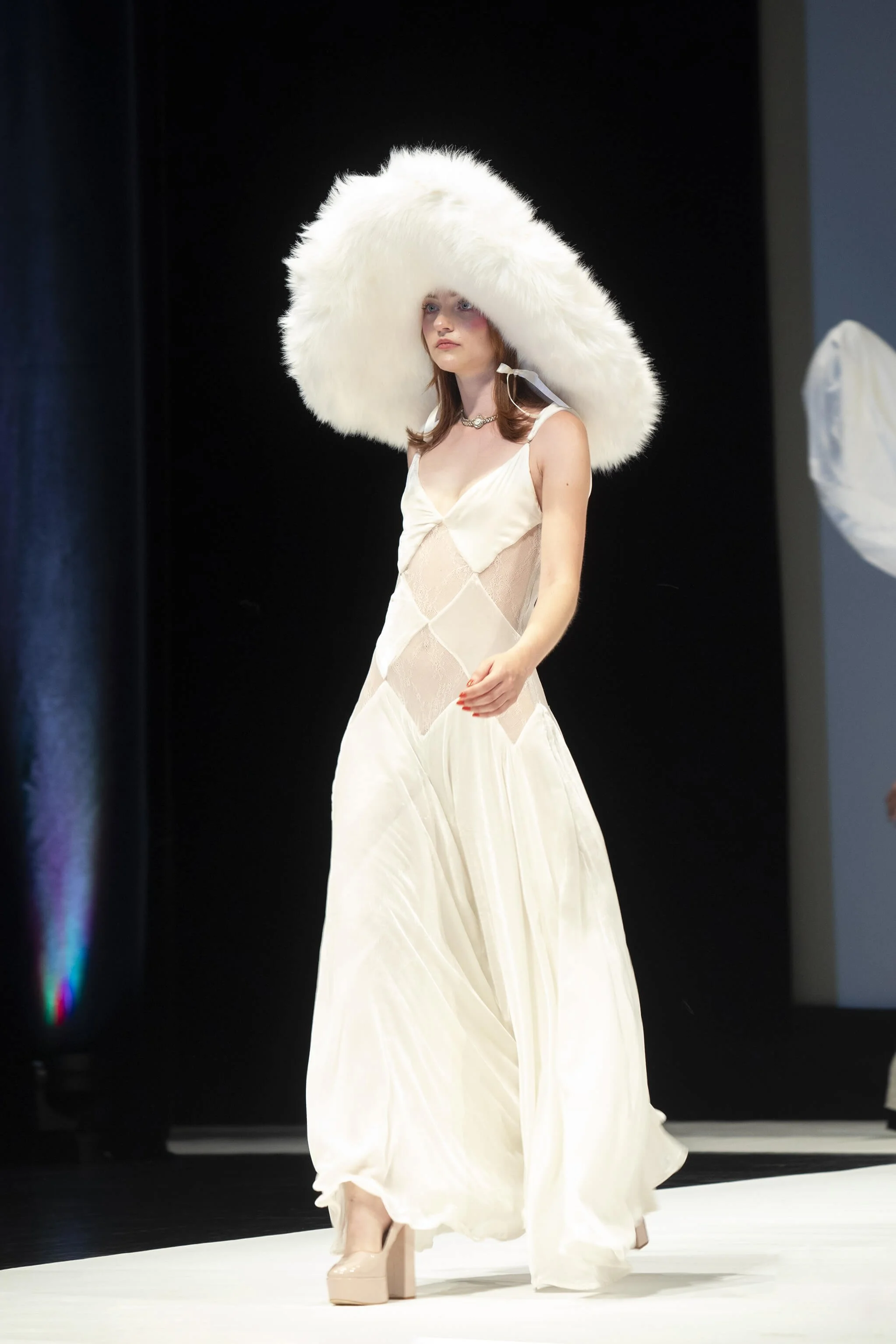 Un modèle élégant défile sur un podium, arborant une robe blanche fluide et un chapeau volumineux en fourrure. La photographie d’événement capture une ambiance fraîche, mettant en valeur le style raffiné du modèle.  Ce cliché reflète le style photogr