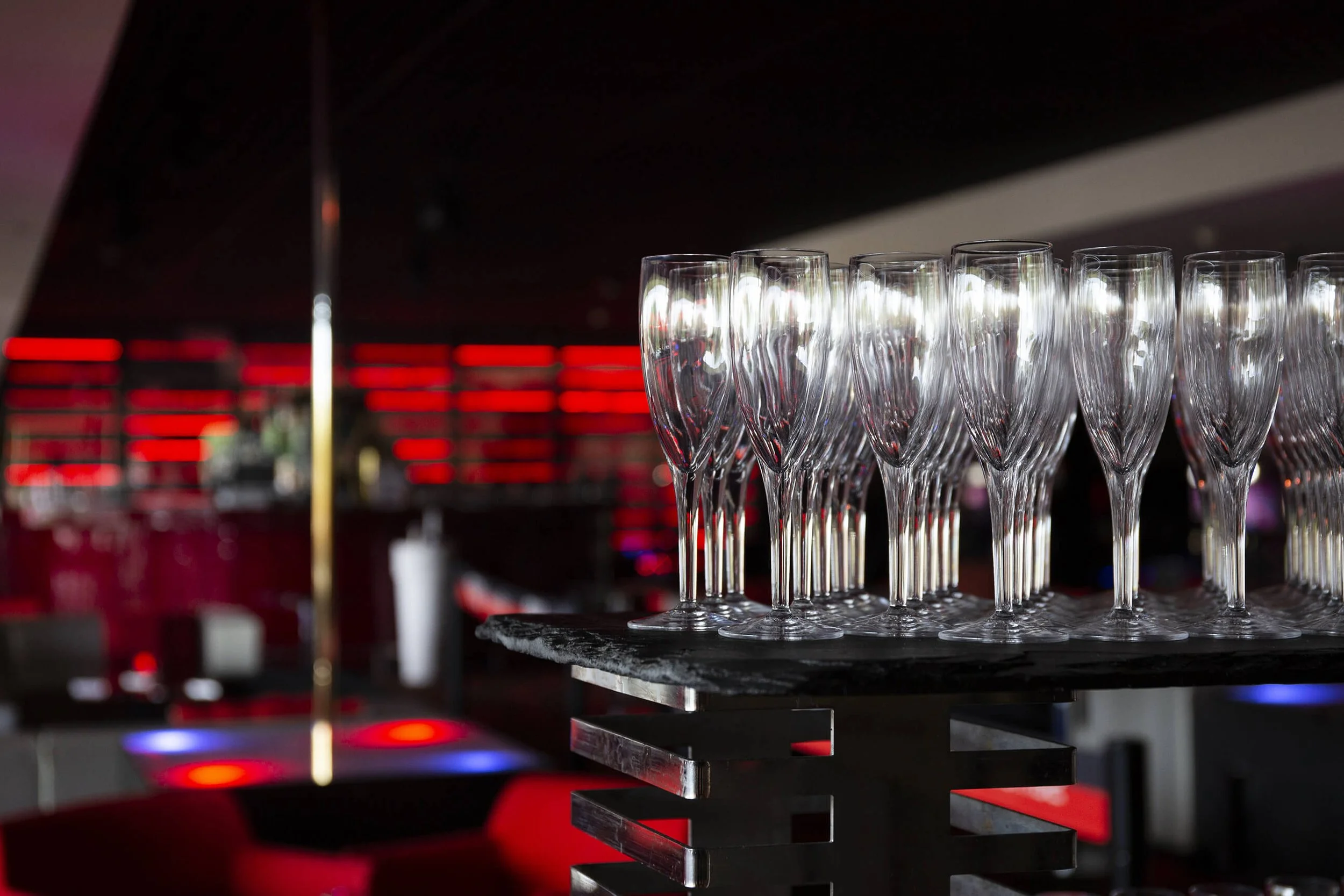 Dans un décor moderne, des verres élégants sont alignés, prêts pour un événement festif. Des lumières rouges et noires créent une atmosphère festive, comme le montre la photographie d’événement professionnelle de François Fauré, qui met en avant le s