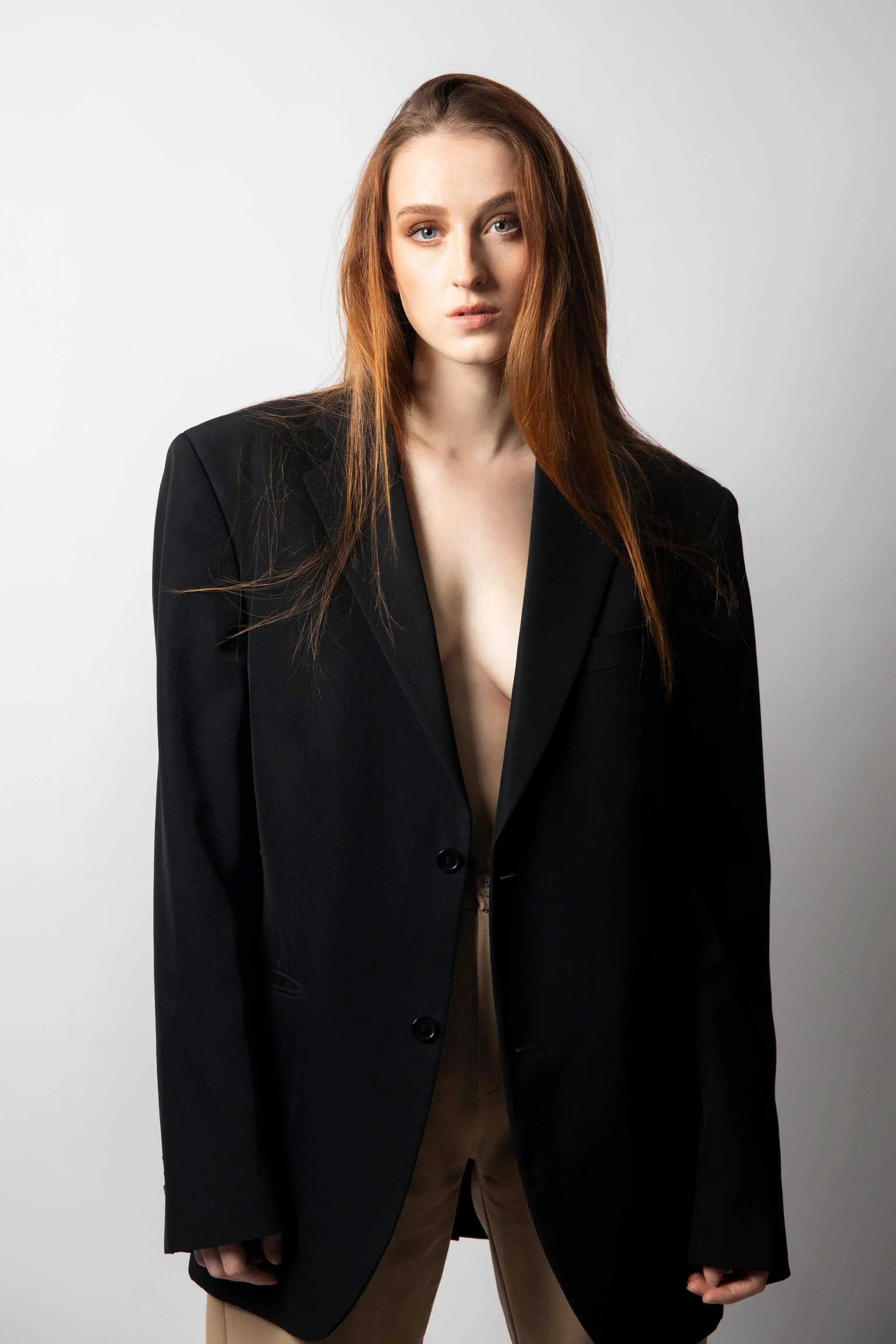 Jeune femme aux cheveux roux et aux yeux bleus portant un blazer noir ouvert en shirt beige, debout contre un fond gris clair.