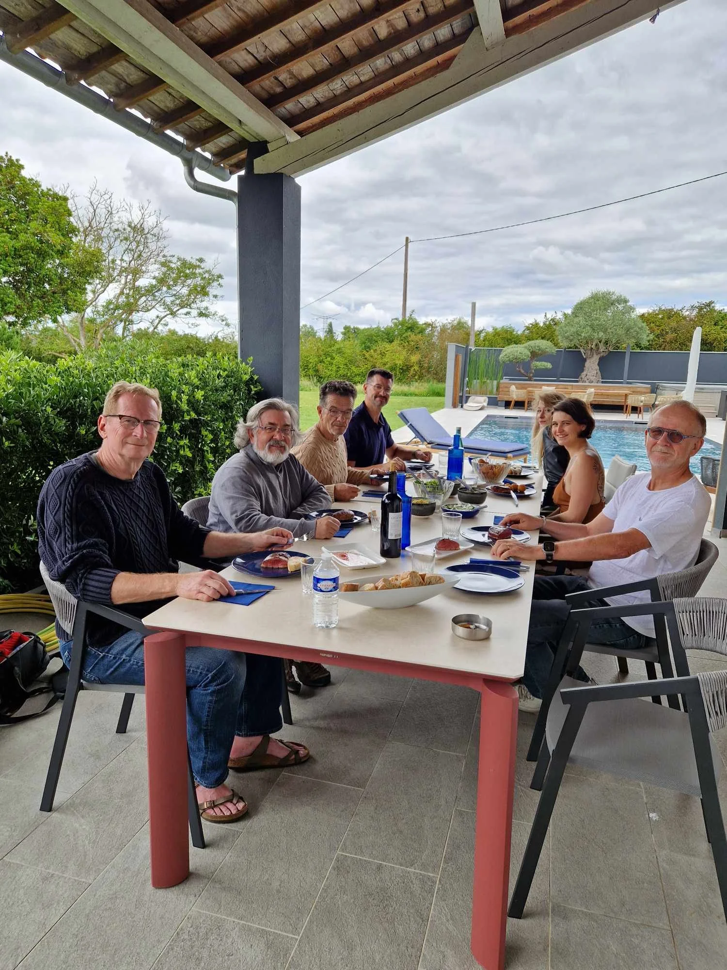 Groupe de sept personnes assises autour d'une table en extérieur, partageant un repas avec une piscine et un jardin en arrière-plan.