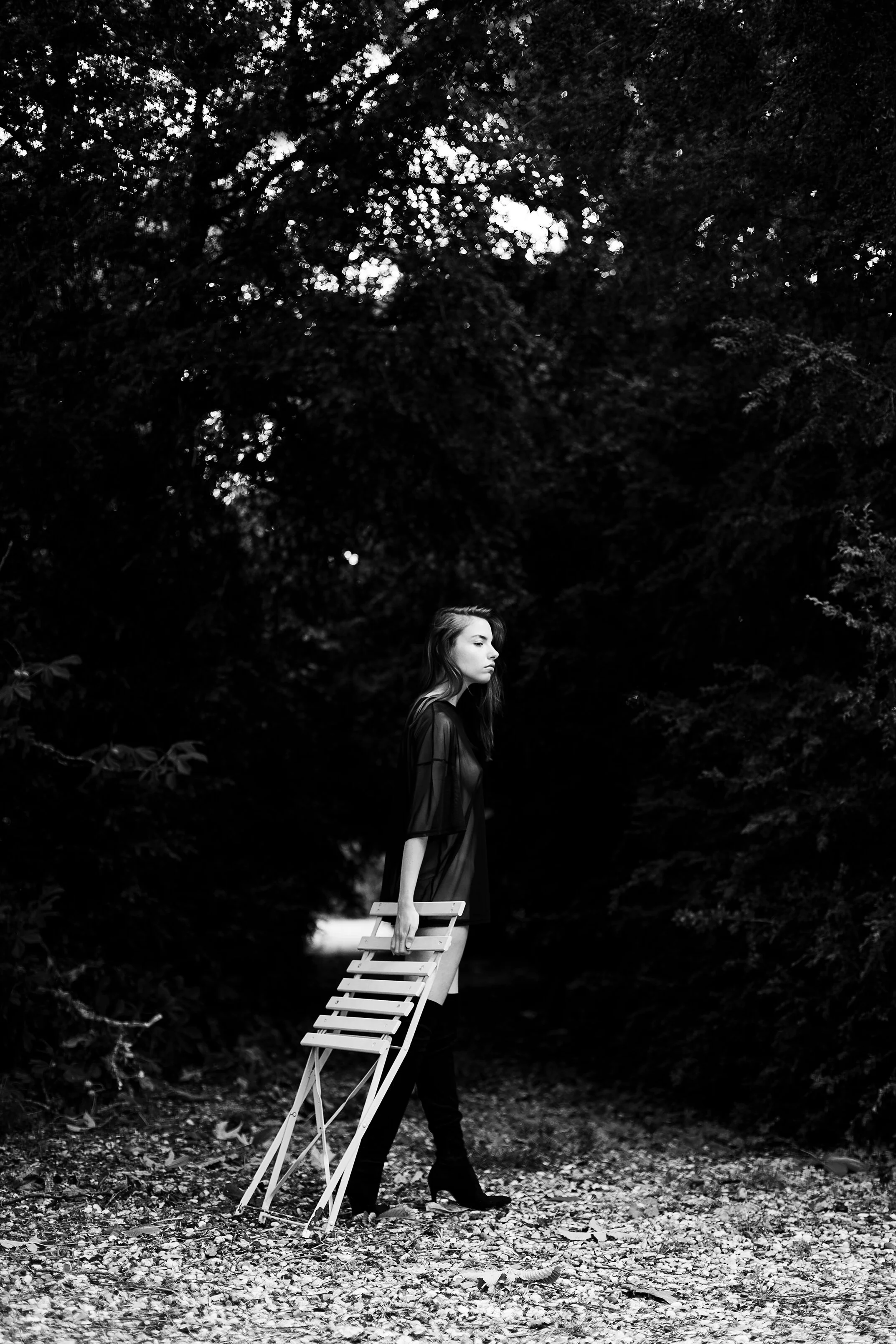 Une femme en vêtements noirs, tenant une chaise pliante, marche dans une forêt sombre, en noir et blanc.