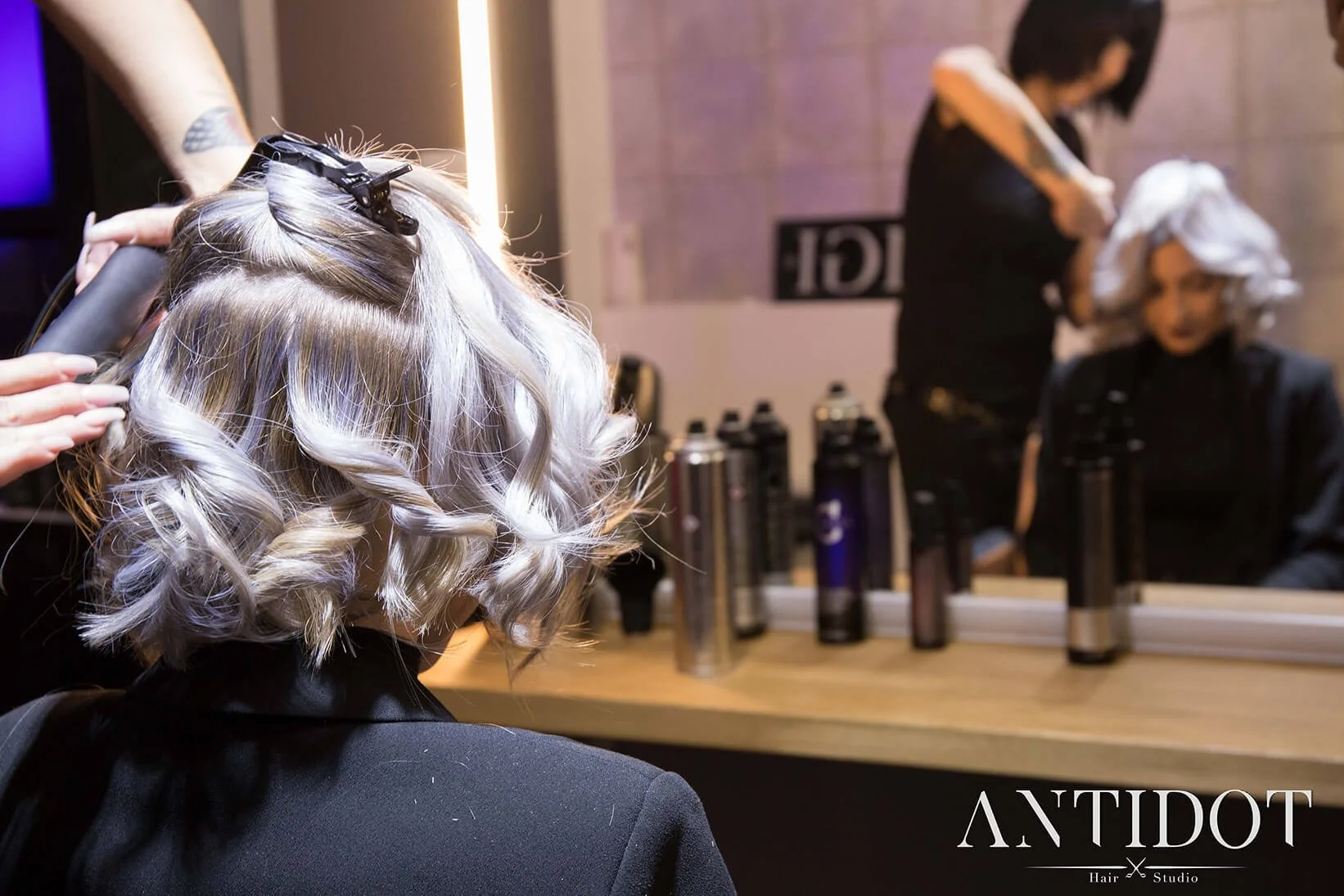 Dans un décor moderne, un professionnel coiffe des cheveux avec soin, entouré de produits de beauté.  L’ambiance est professionnelle, avec des stylistes au travail et des produits en arrière-plan. Une séance de coiffure professionnelle met en avant l