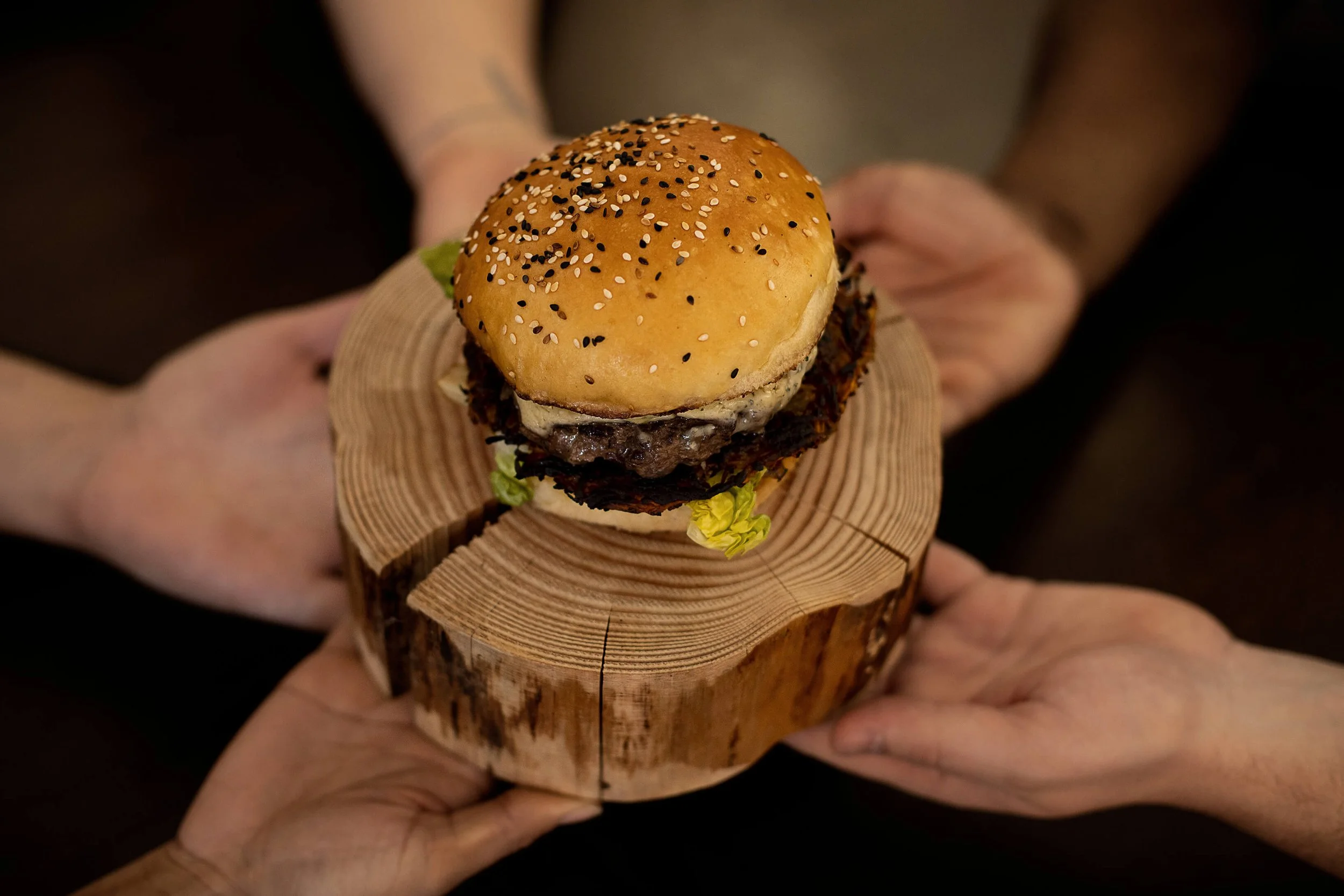 Un hamburger artisanal est présenté sur un support en bois, tandis qu’un hamburger gourmet est montré avec des mains en arrière-plan. Un burger frais, parsemé de graines de sésame, repose sur un plateau en bois.  Plusieurs personnes partagent un hamb
