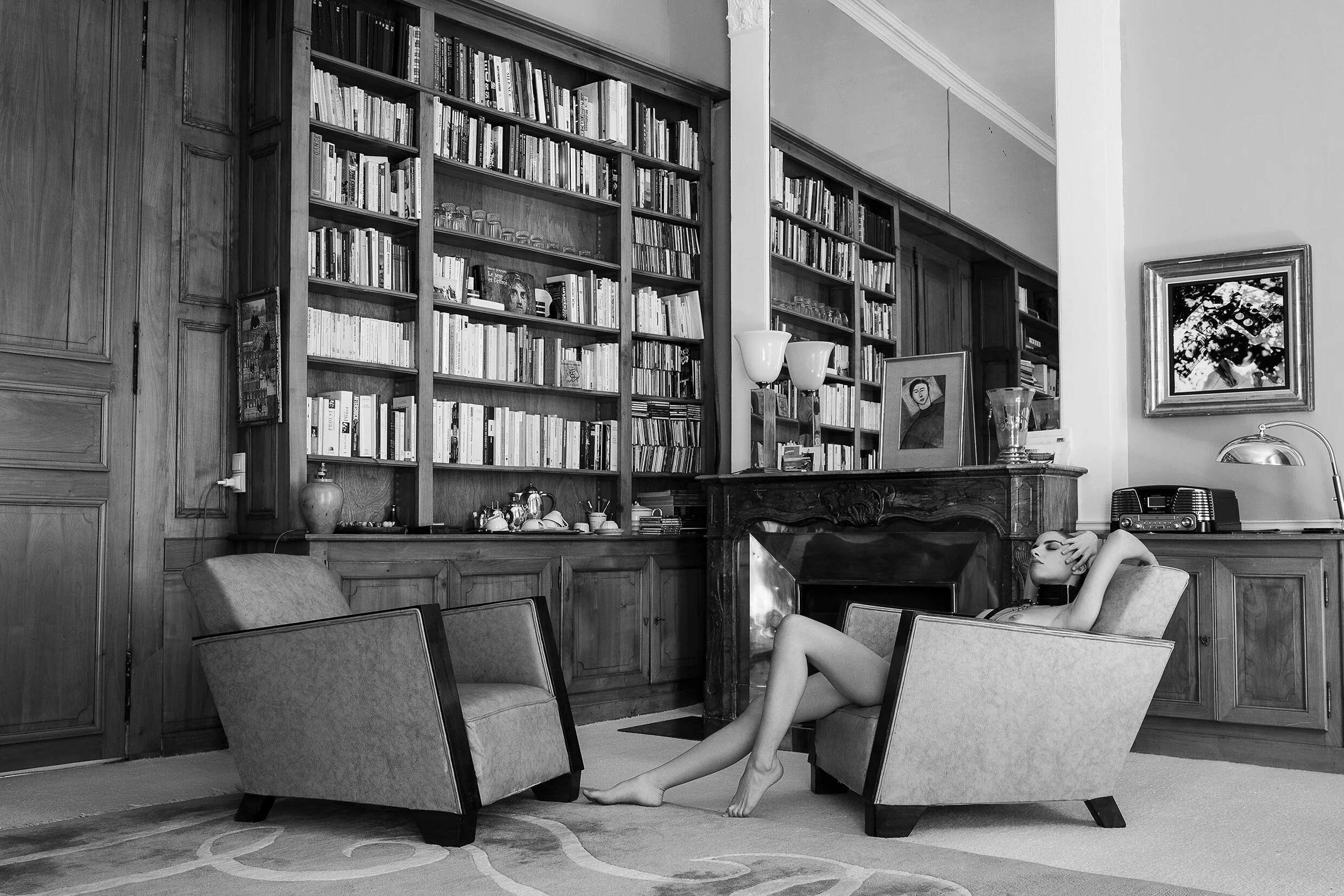 Une femme nue portant un masque de visage, assise dans un salon avec une grande bibliothèque en bois, un feu de cheminée, des fauteuils, et des œuvres d'art. Parapluie sur la bibliothèque.
