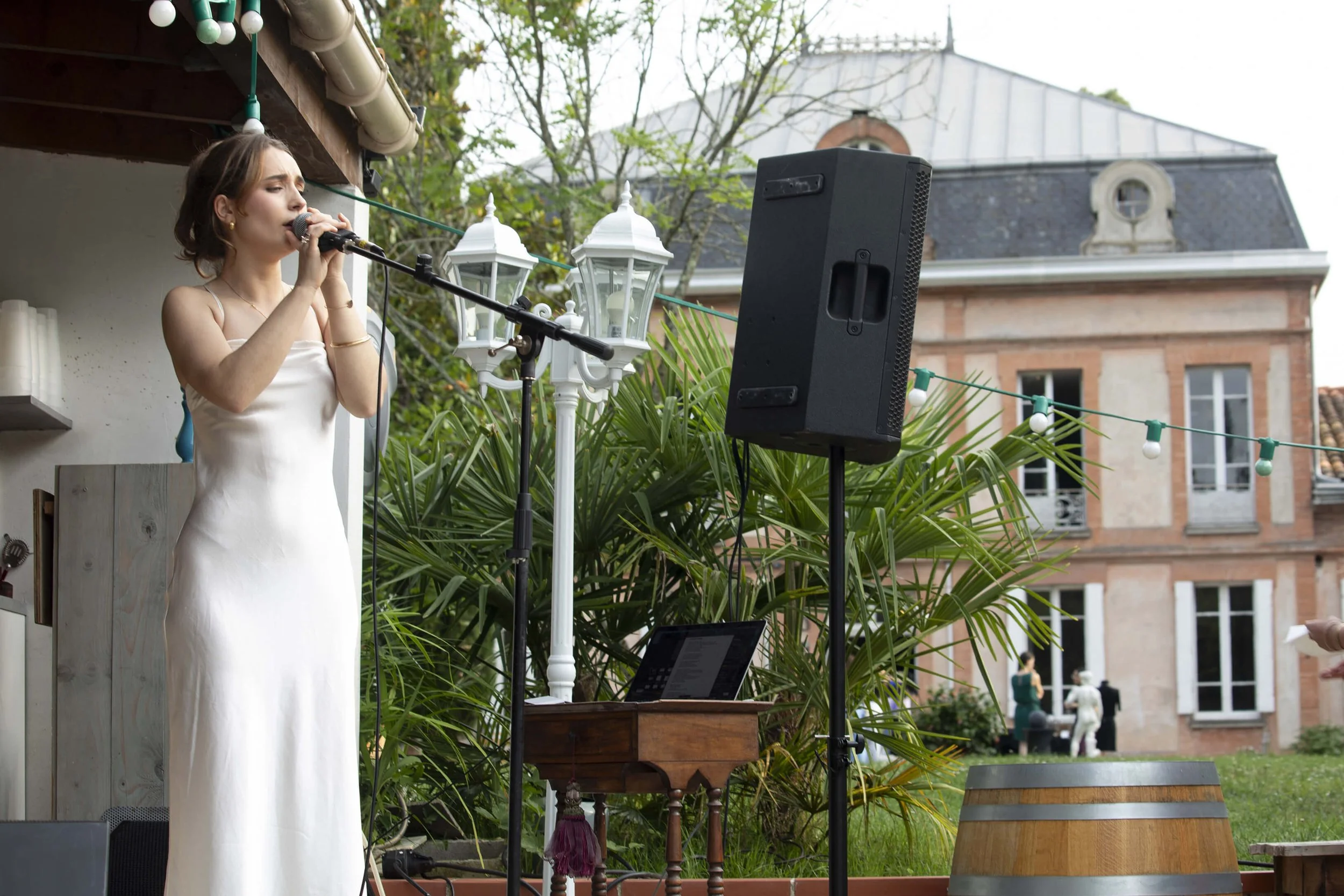 Une chanteuse vêtue d’une robe blanche se produit lors d’un événement en plein air, entourée d’un décor verdoyant.  L’ambiance est élégante, avec des lampions illuminant l’espace.  Le photographe François Fauré immortalise la performance musicale.  L