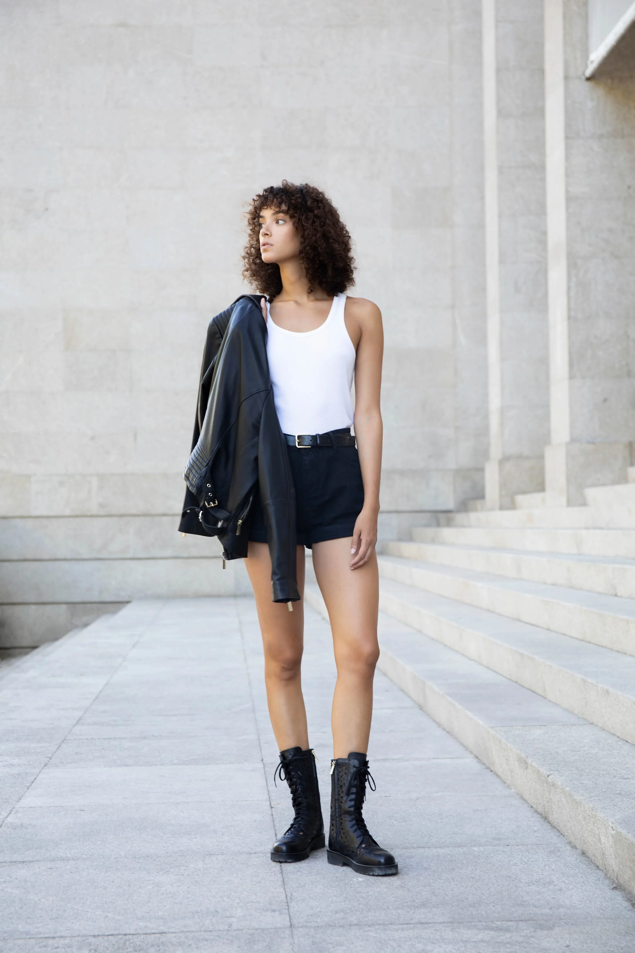 Jeune femme avec veste en cuir noire sur l'épaule, debout devant un mur en pierre, portant un débardeur blanc, short noir et bottes noires.