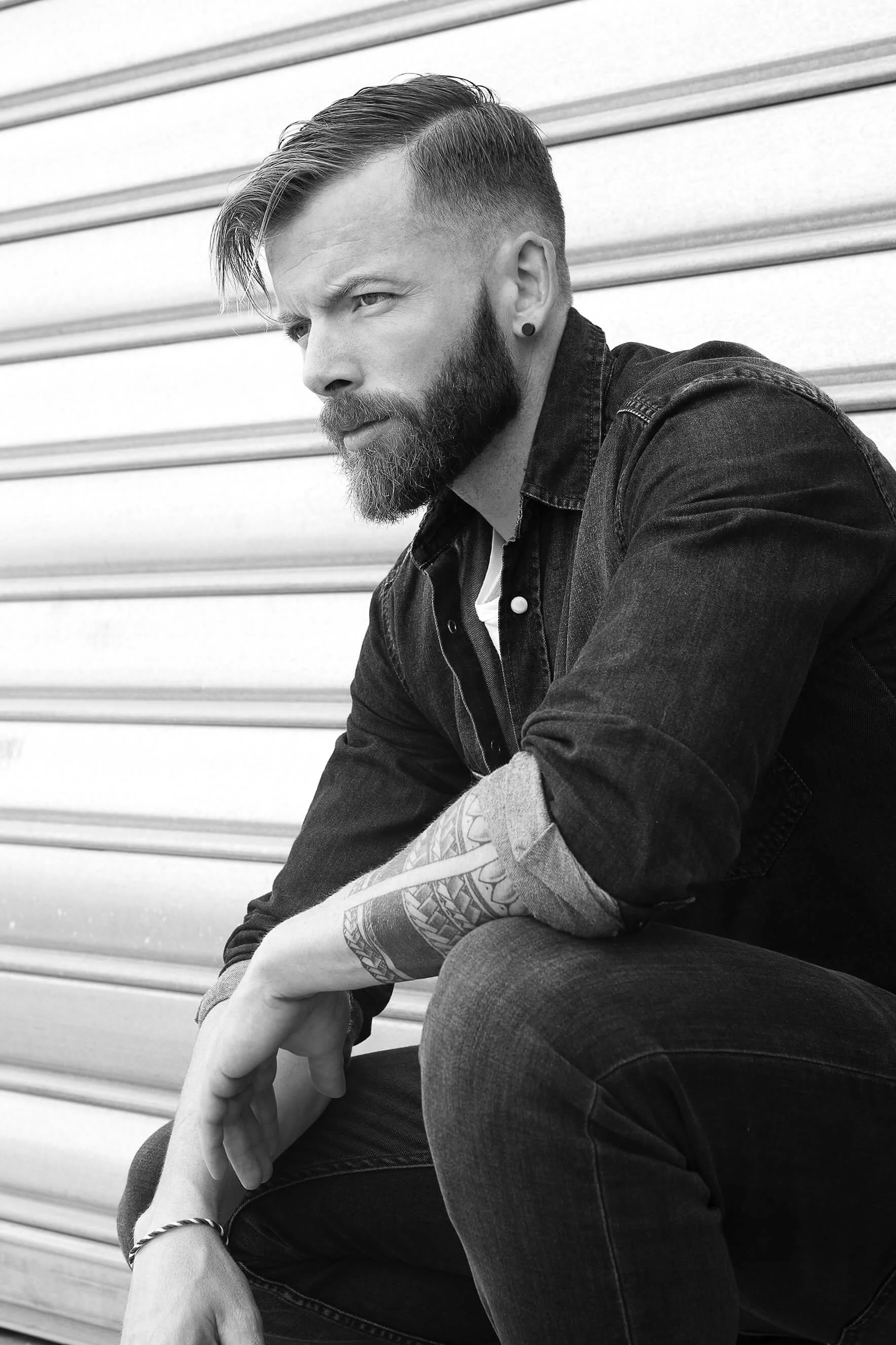 Homme avec barbe et tatouage en noir et blanc, assis contre un mur en métal