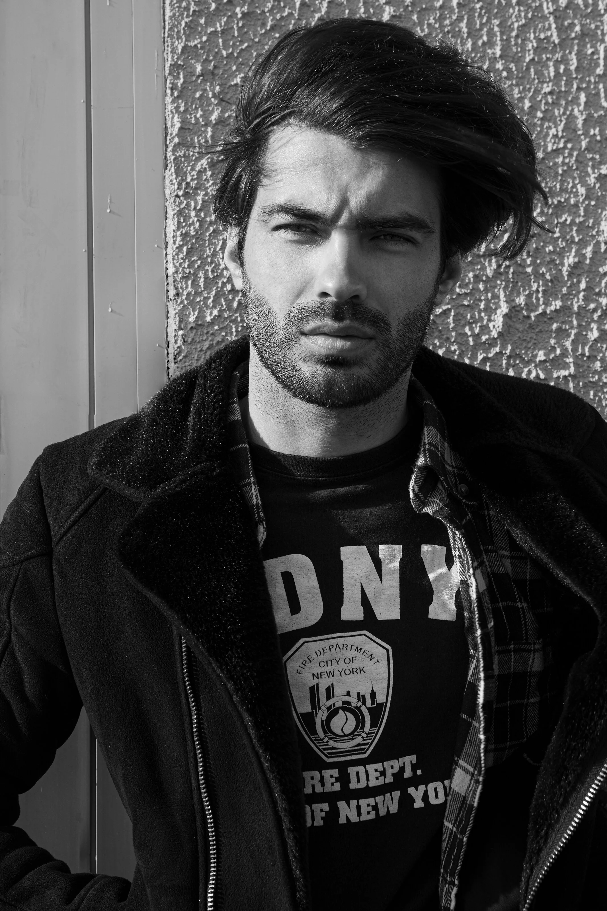Un homme avec des cheveux longs et une barbe, portant un manteau noir et un t-shirt avec un logo de la ville de New York, posé devant un mur en béton. L'image est en noir et blanc.