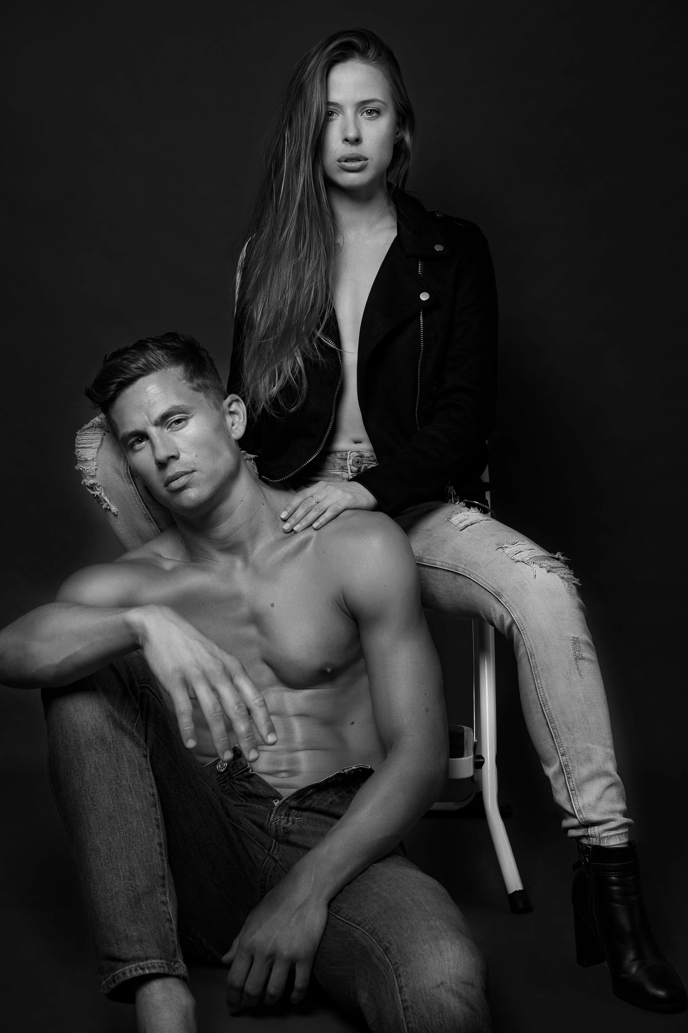 Une photographie en noir et blanc de deux personnes, une femme debout et un homme assis, tous deux avec une allure expressive et un style moderne, dans un studio sombre.