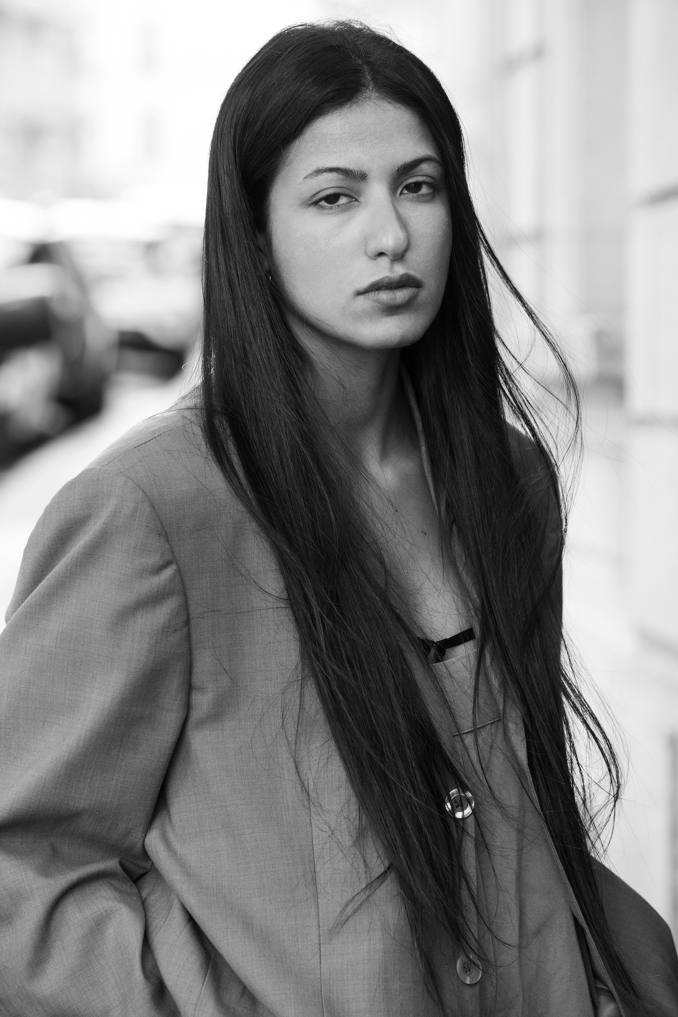 Photo en noir et blanc d'une jeune femme avec des cheveux longs, porte un blazer et regarde l'objectif, fond flou d'une rue urbaine.