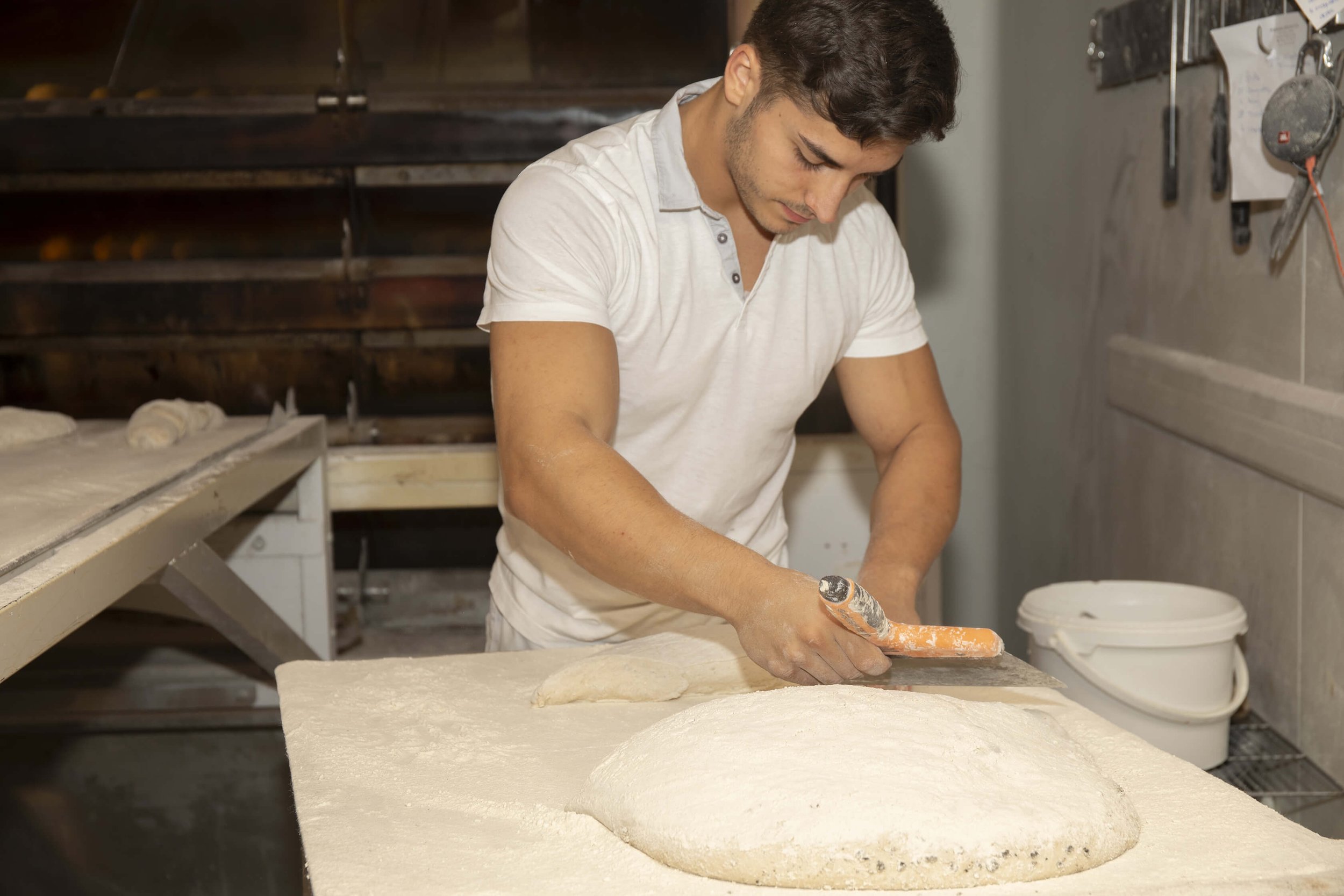 Dans une boulangerie professionnelle, un boulanger façonne la pâte avec expertise.  La préparation artisanale du pain met en avant l’utilisation d’ingrédients frais. L’ambiance chaleureuse de la boulangerie est accentuée par la présence du boulanger 