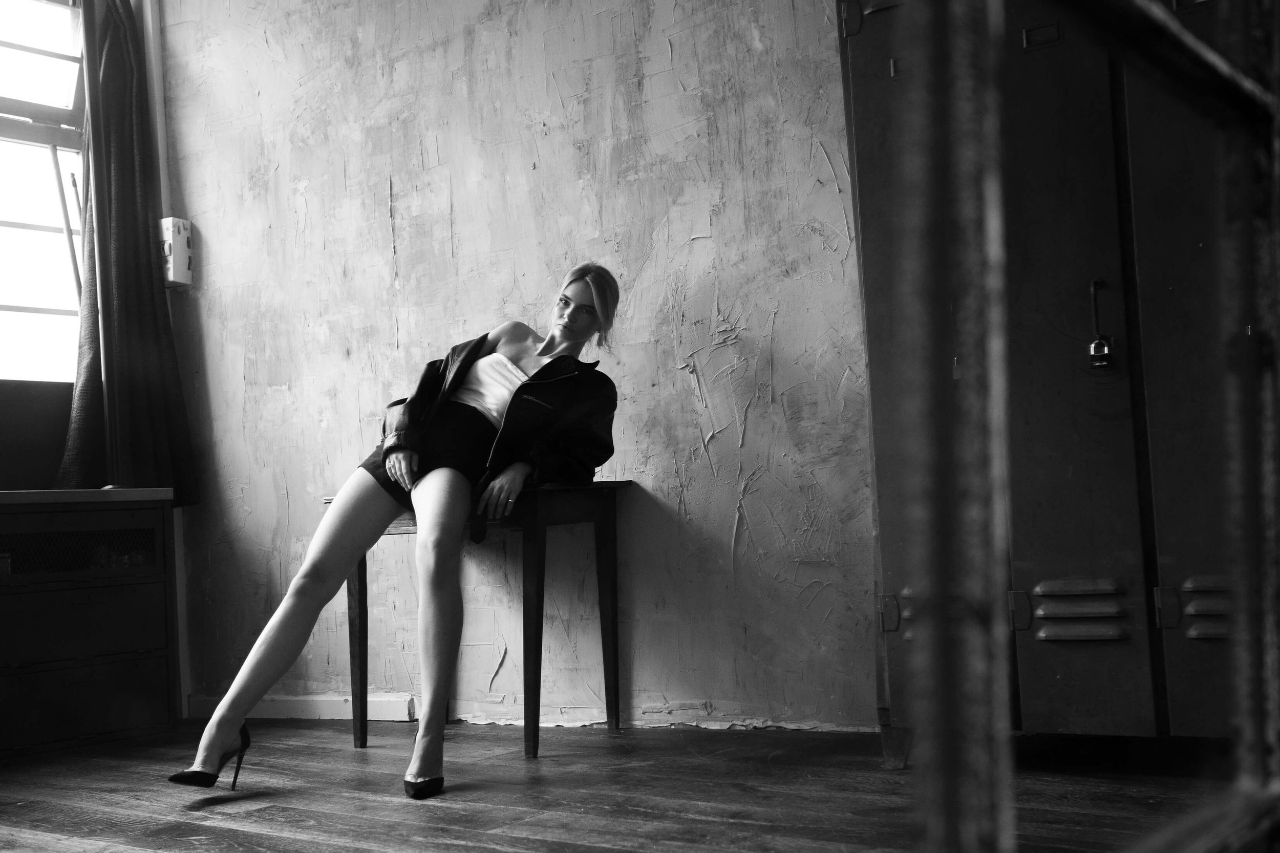 Une femme posant de manière artistique dans une pièce sombre avec une lumière naturelle venant d'une fenêtre, portant des talons, un t-shirt blanc et une veste noire.